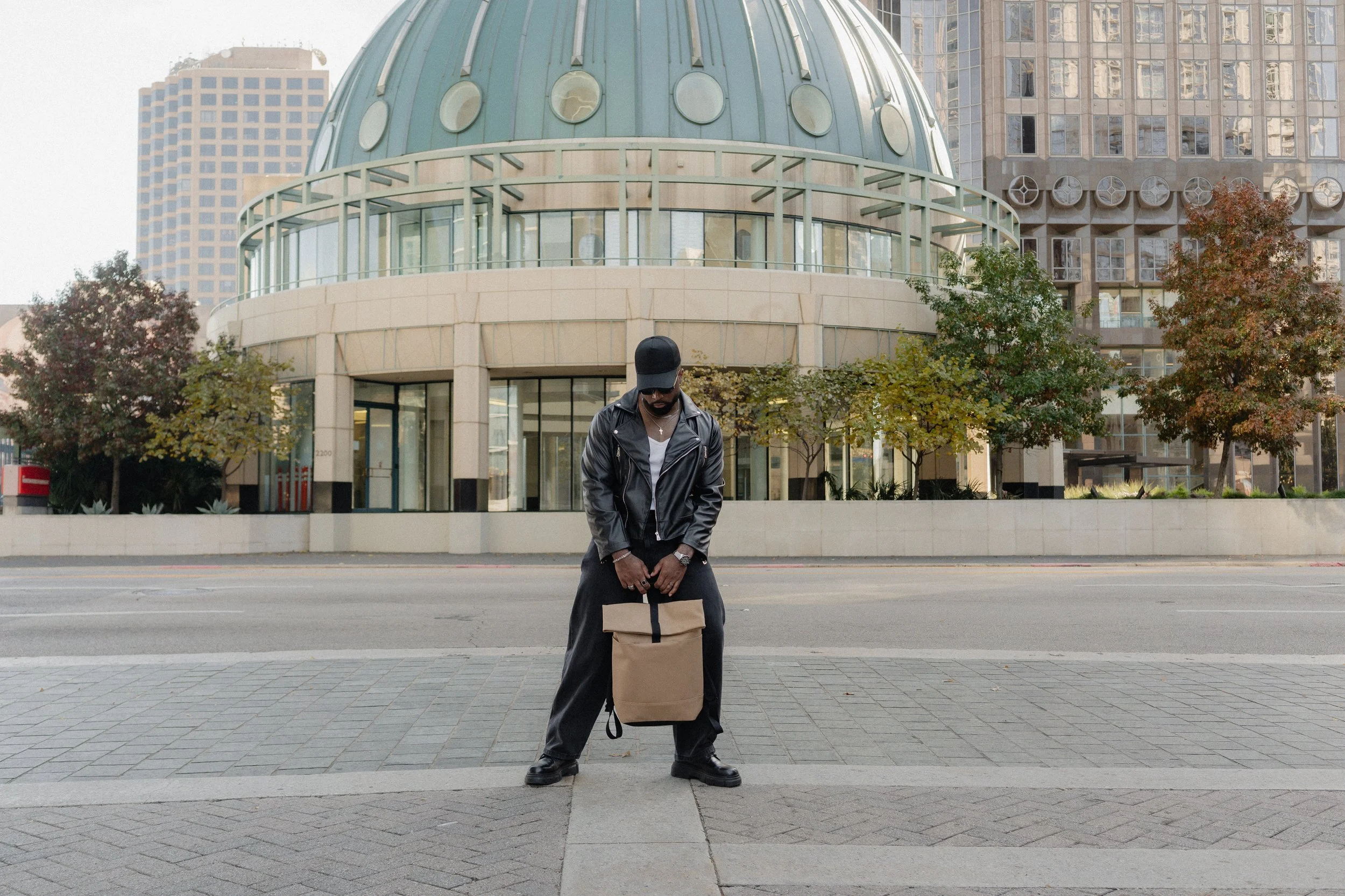 Man dressed in black leather jacket, black pants, black cap, and black shoes standing on sidewalk in front of a futuristic building with glass windows and trees, holding a brown designer backpack.