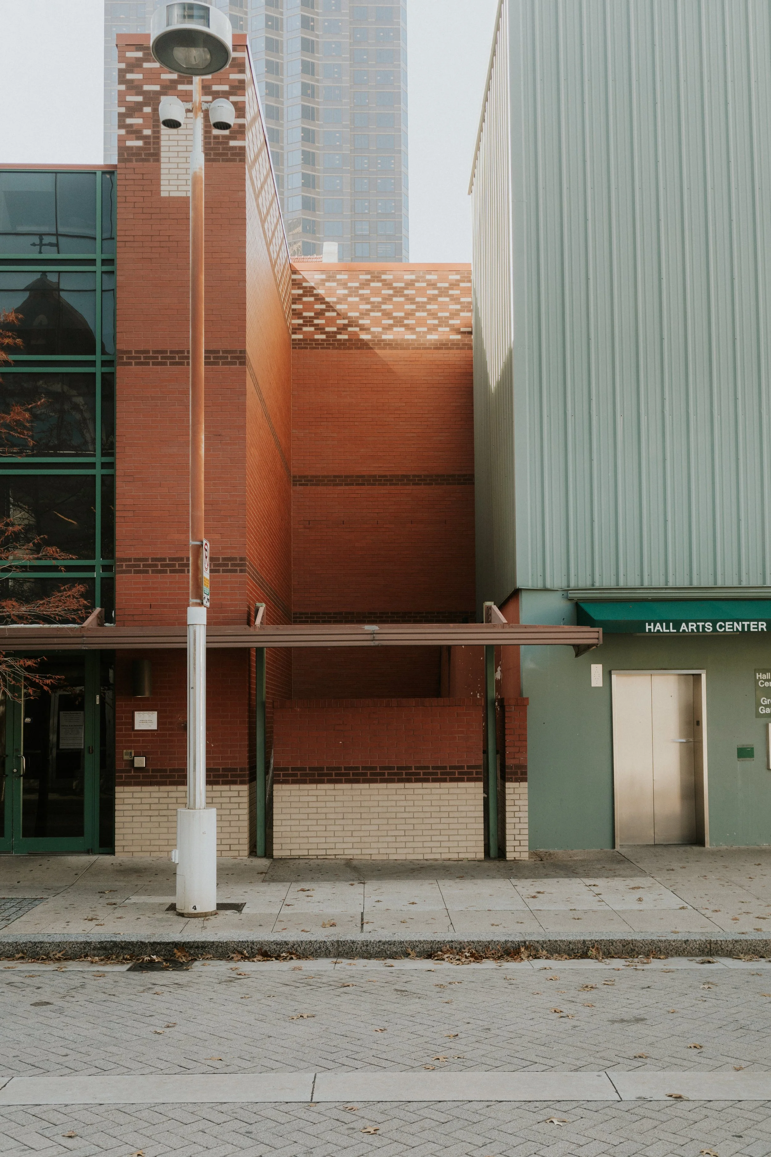An urban scene showing a building with red brick walls and a glass section, a metal-sided structure, a streetlamp in the foreground, and a building with a sign that reads 'Hall Arts Center'.