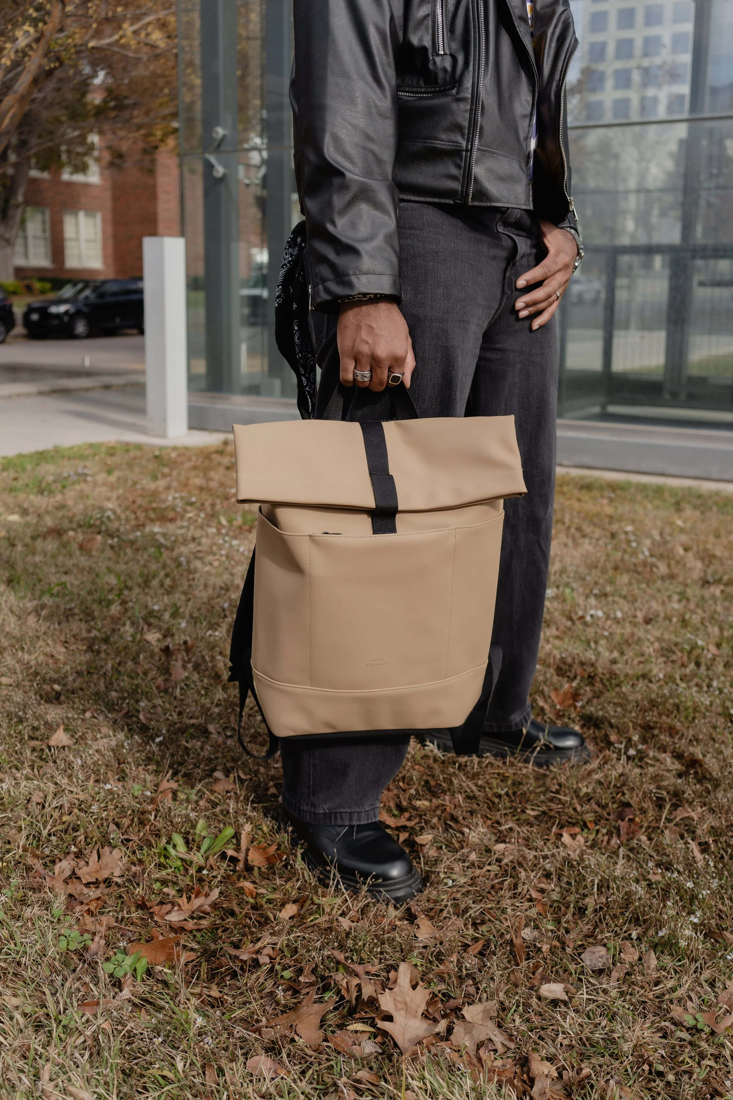 Person standing outdoors holding a beige backpack with black straps, wearing dark jeans, black shoes, and a black leather jacket.