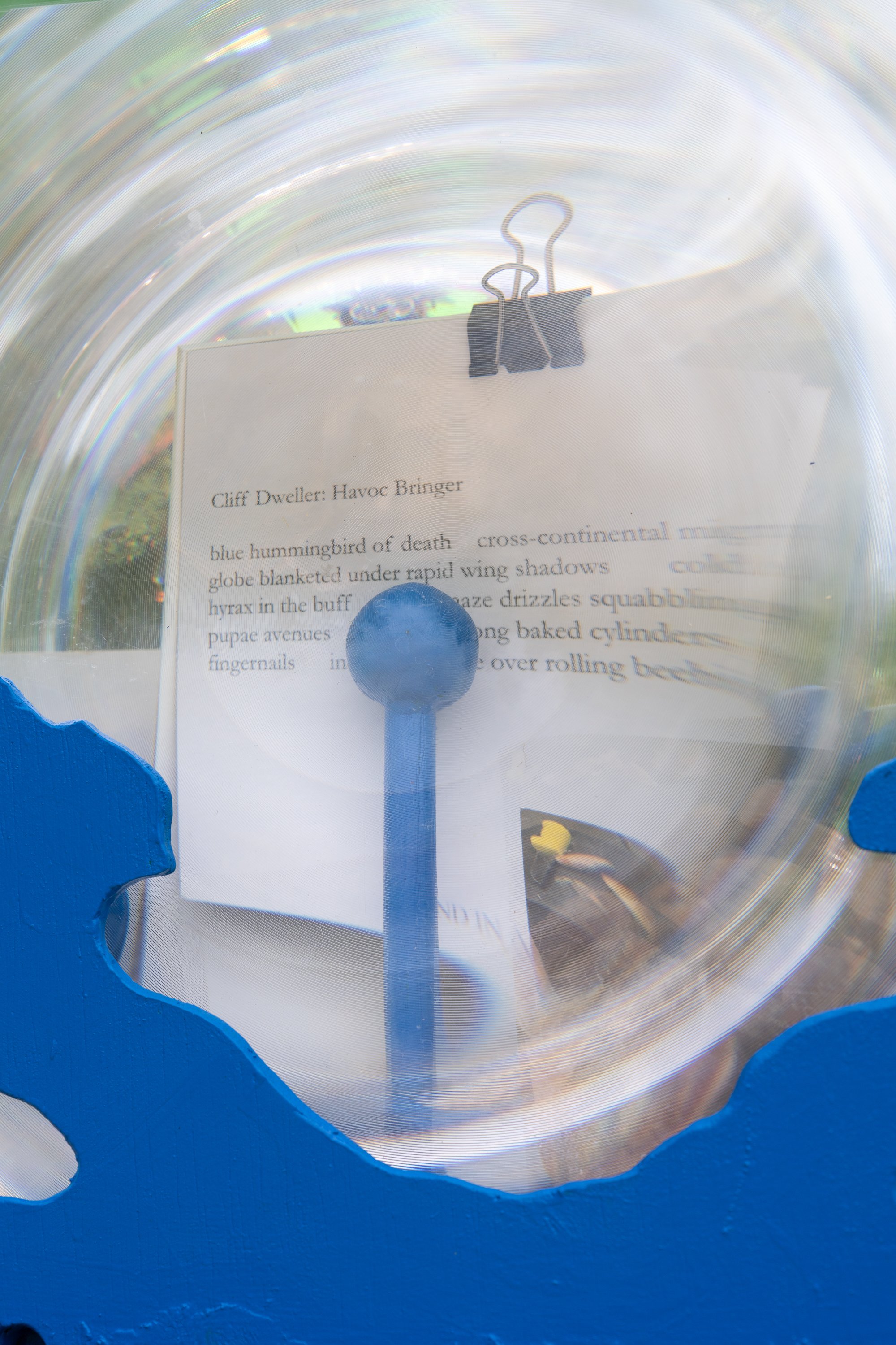 A poem on postcard distorded under plexiglass held by blue sculpture.