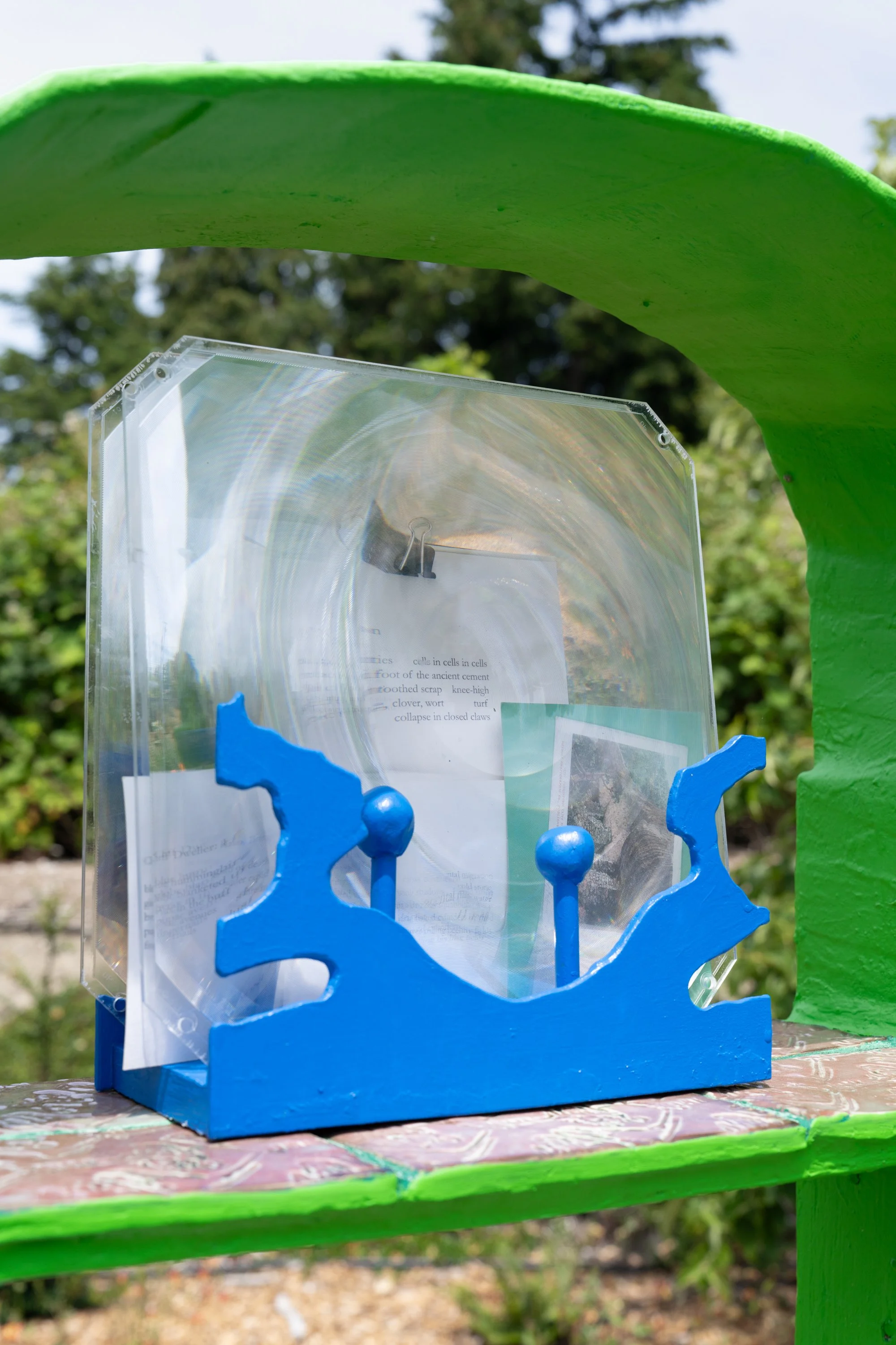 A poem on postcard distorted under plexiglass held by blue sculpture.