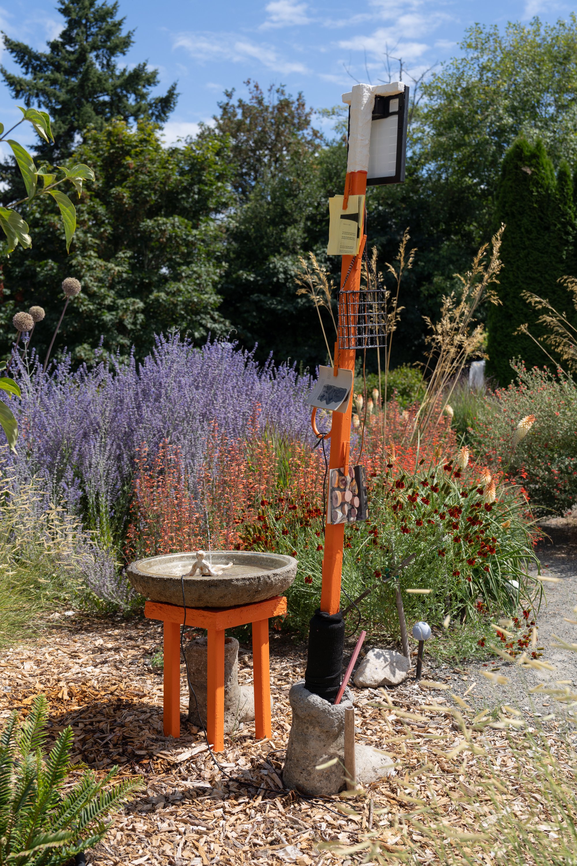 An orange sculpture with a fountain in a colorful garden.