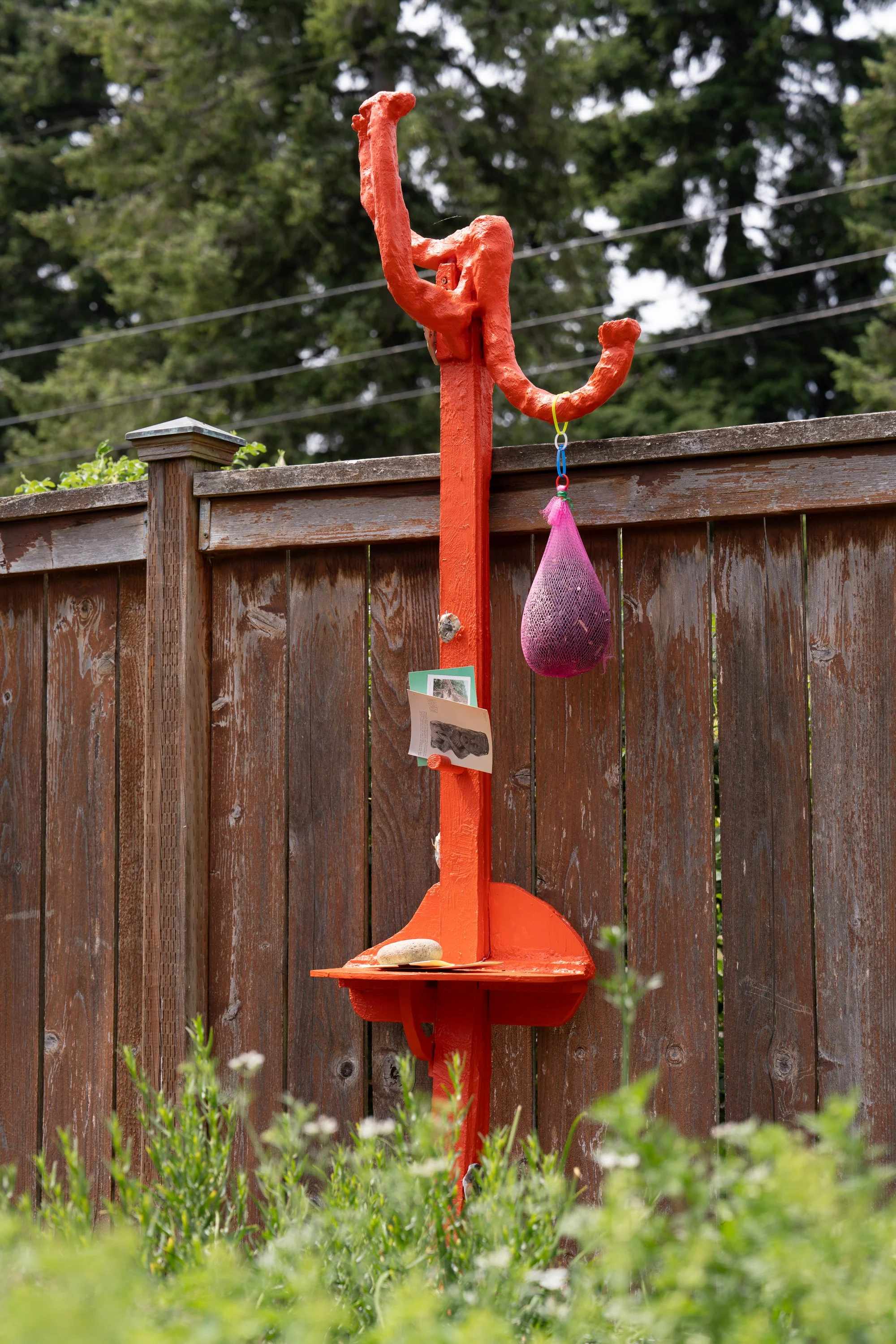 An orange sculpture leaning against a garden fence.