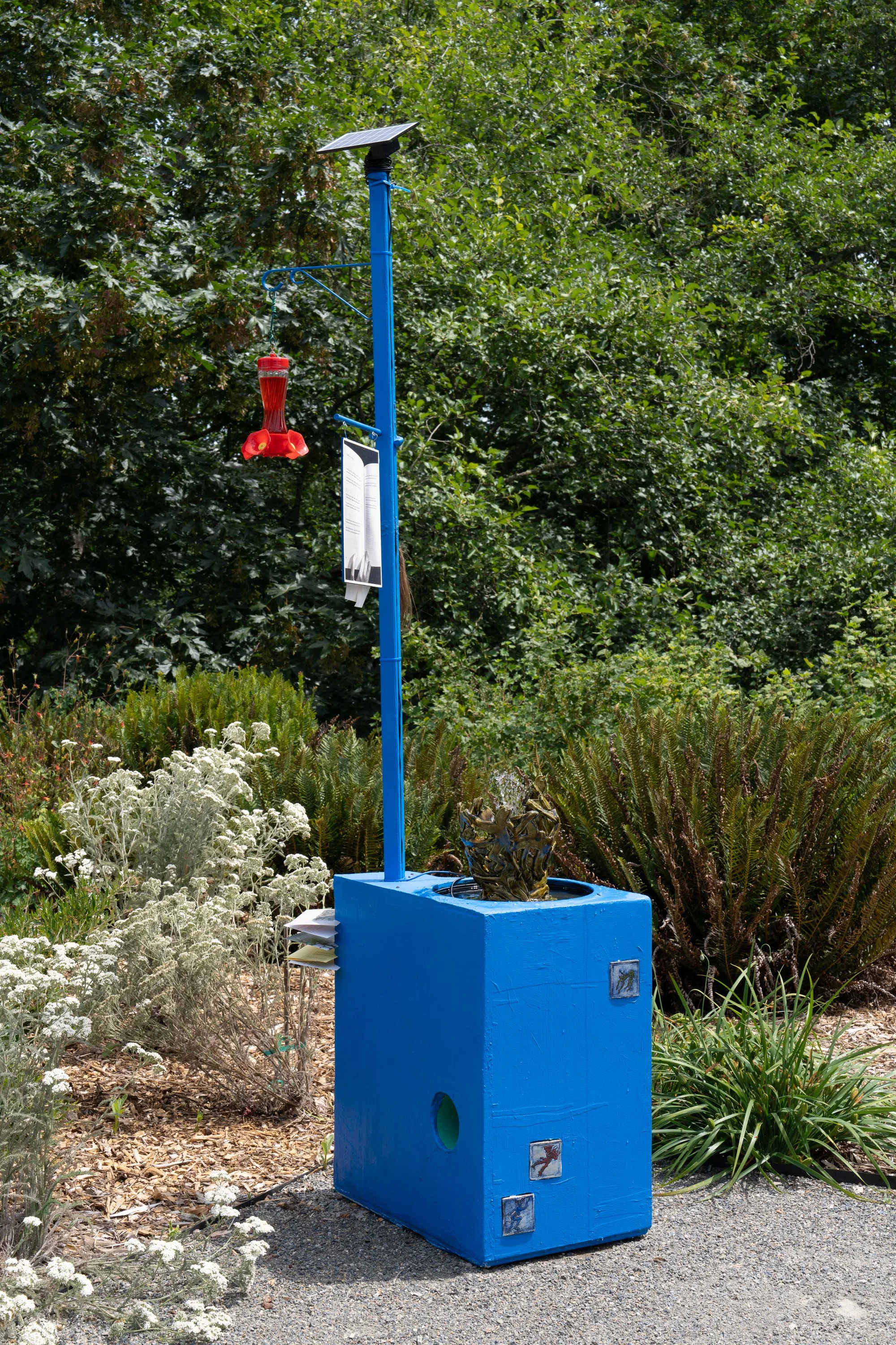 Blue sculpture with red humming bird feeder in a garden.