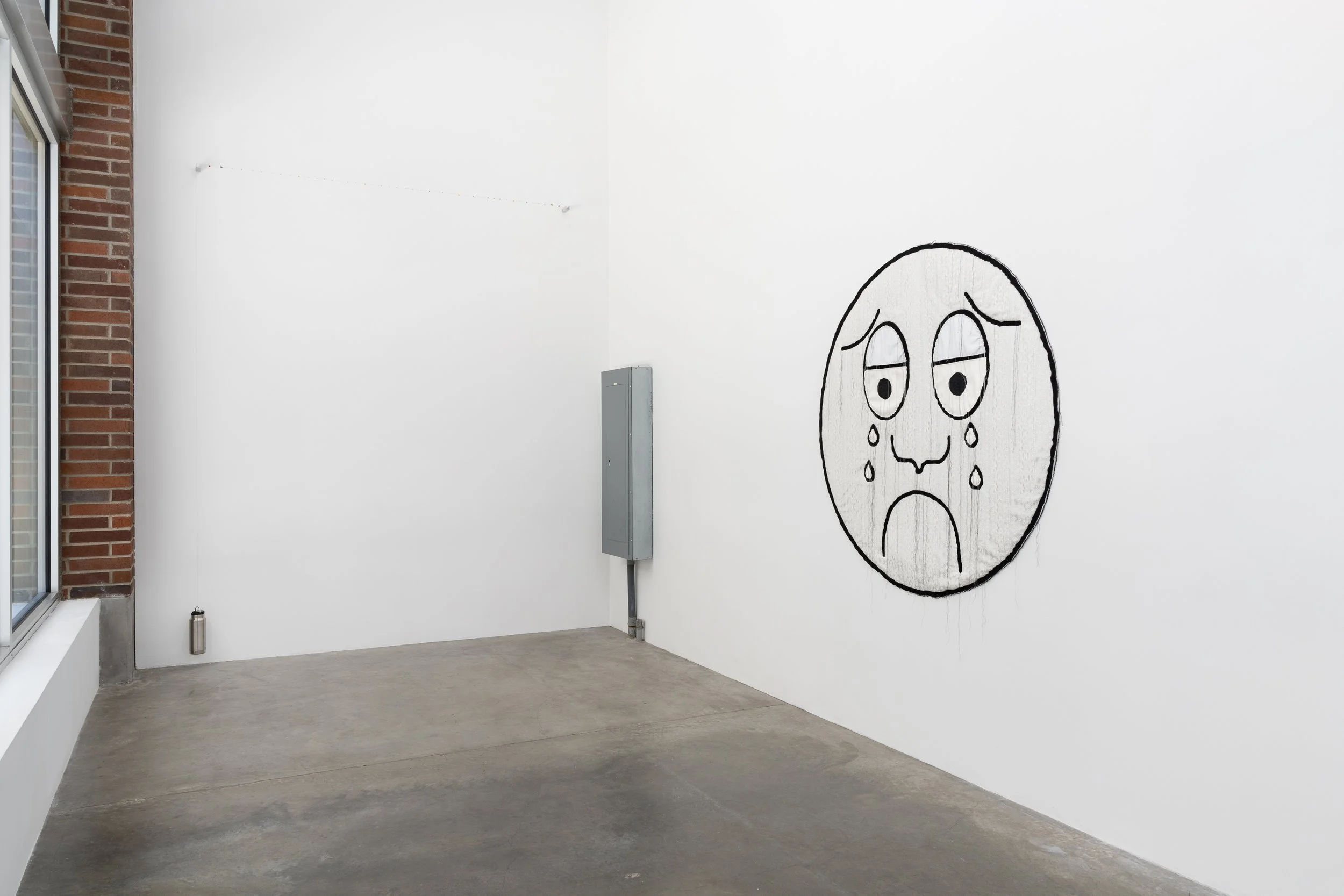 A crying face made of textile and a string of small objetcs on the walls of a gallery.