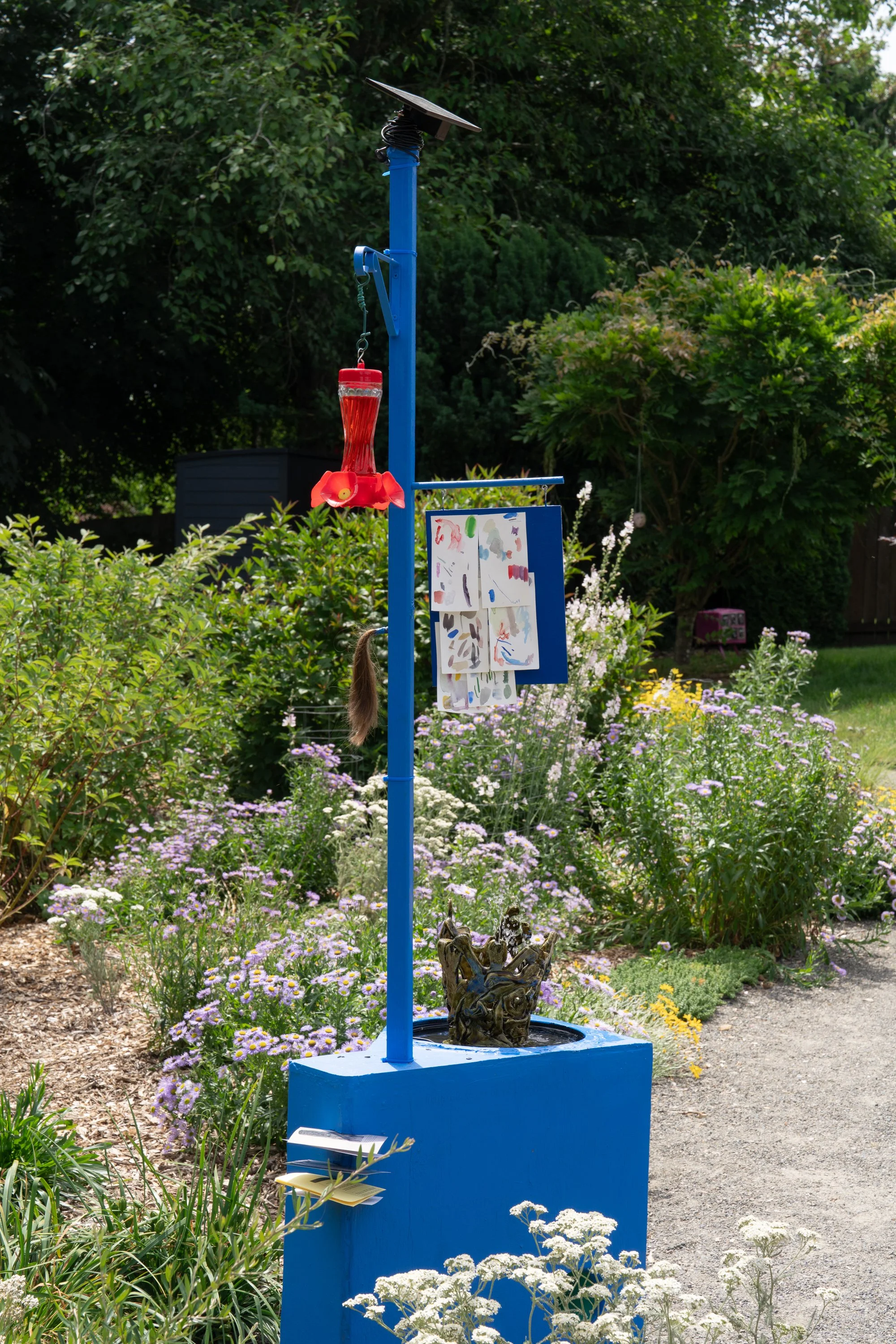 Blue sculpture with red humming bird feeder and watercolor paintings in a garden.