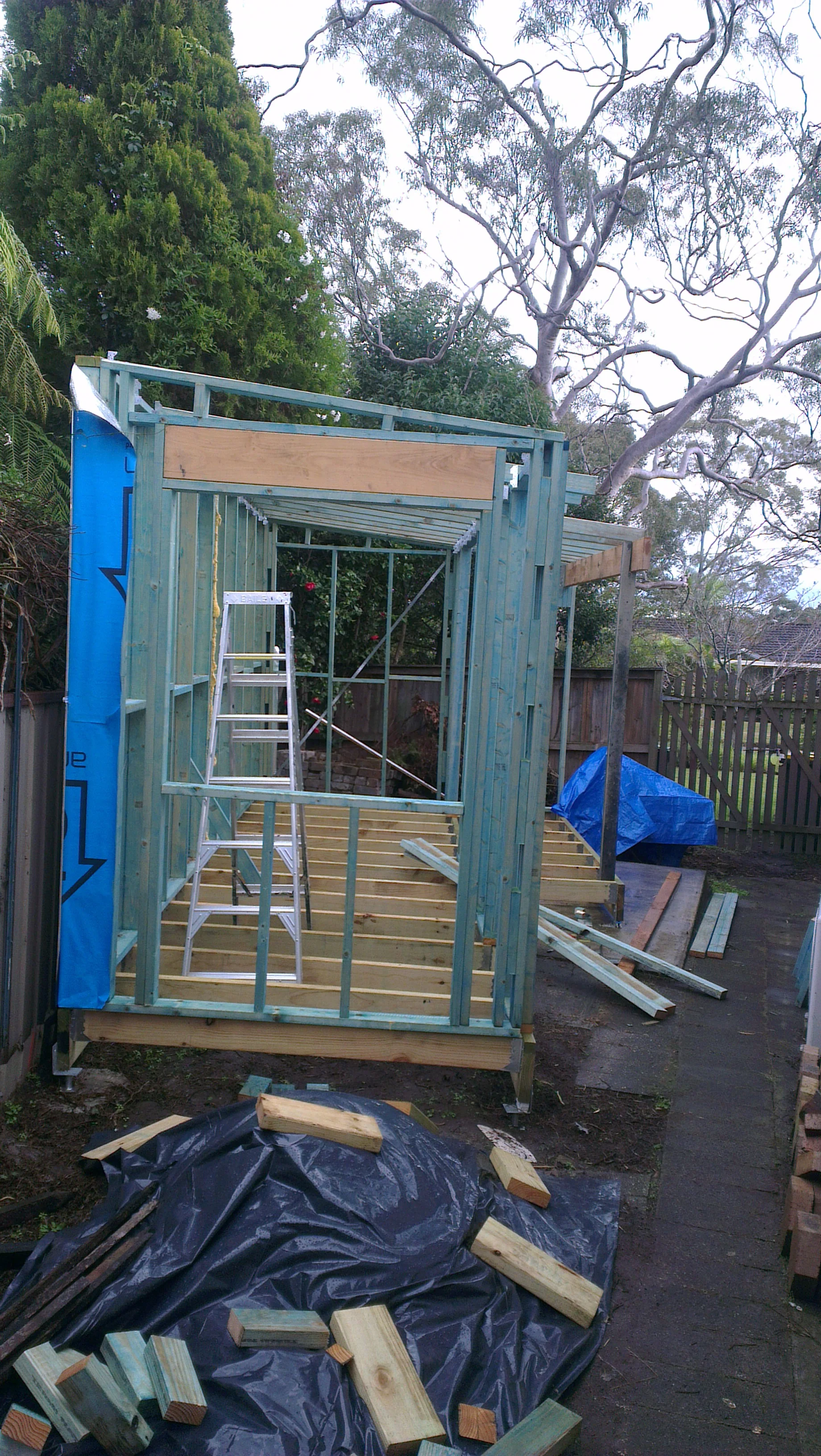 A shed being built, floor joists and wall supports can be seen with an aluminium ladder inside. More building materials are on the ground.