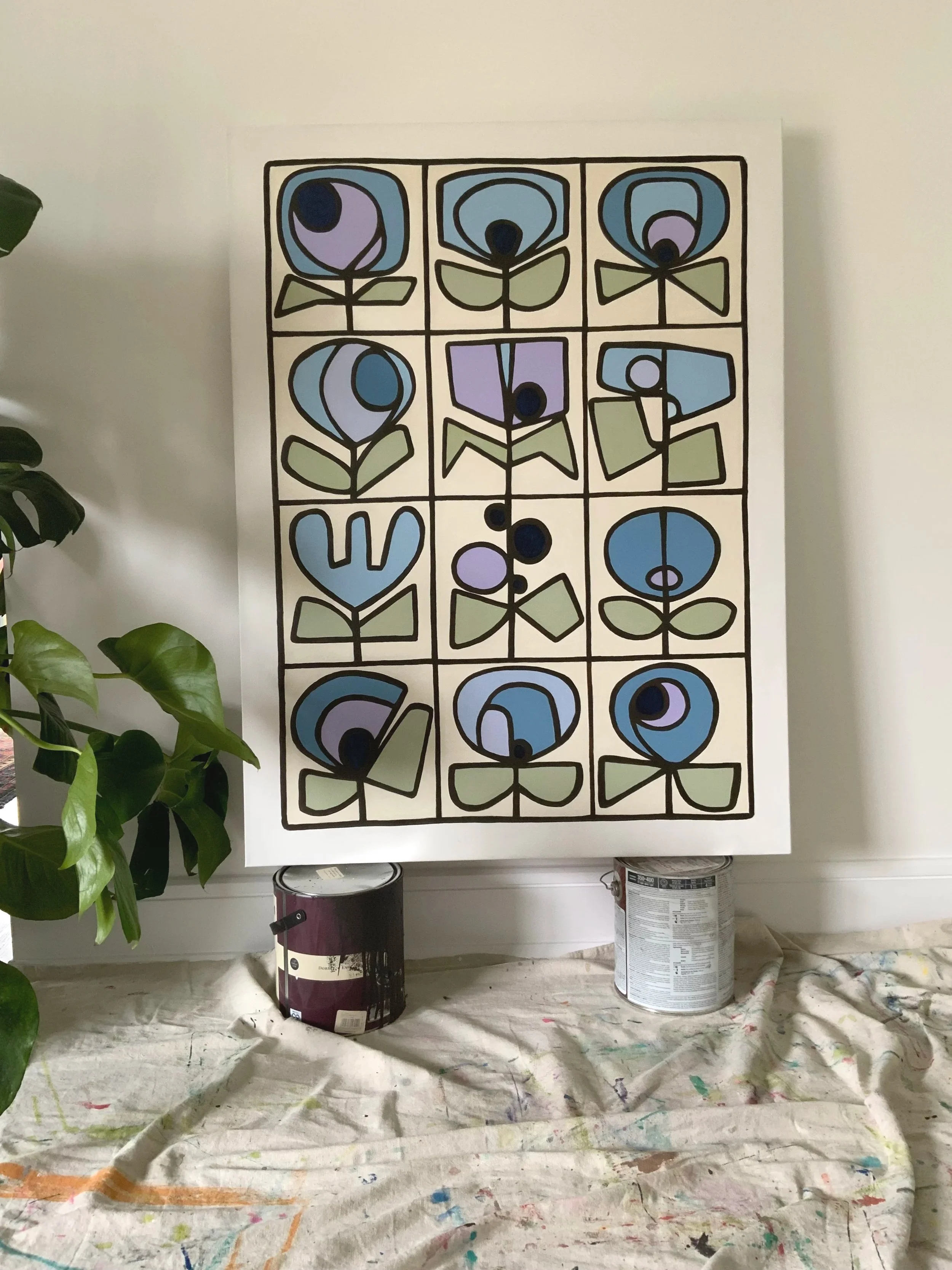 A colorful geometric floral painting on a large vertical canvas, surrounded by paint cans on a painter's cloth, with green leaves from a nearby plant visible on the left. Denise Mortensen is a New Jersey abstract artist known for SHELFIES® — colorful