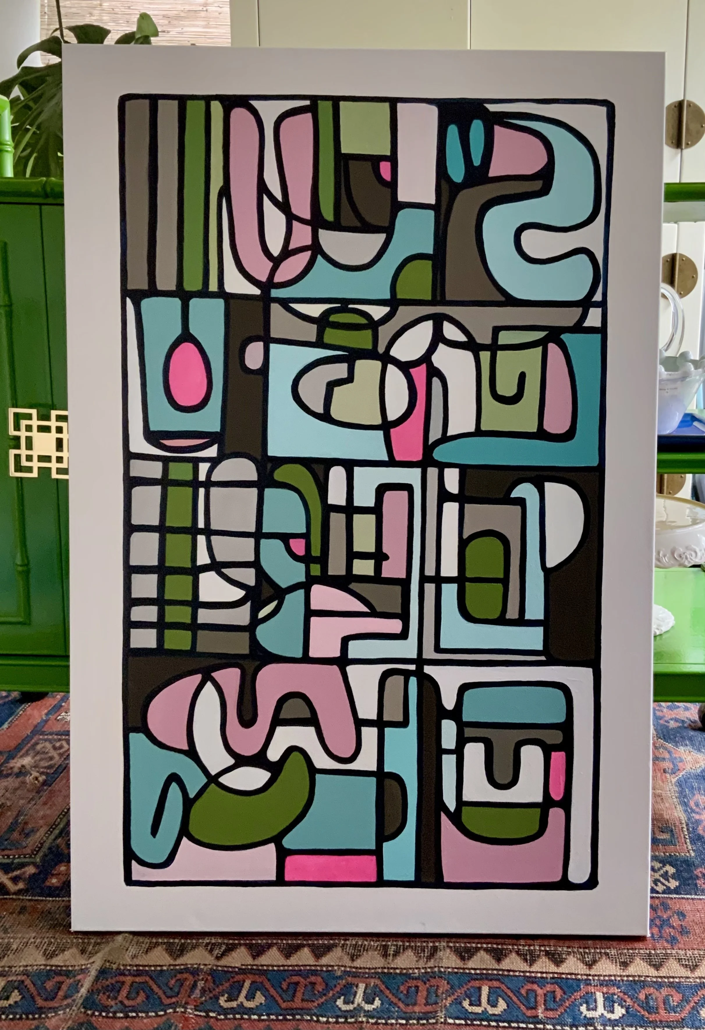Abstract geometric painting with various shapes and colors, including pink, blue, green, brown, and white, outlined in black, mounted on a white panel. Denise Mortensen is a New Jersey abstract artist known for SHELFIES® — colorful, bright, and senti