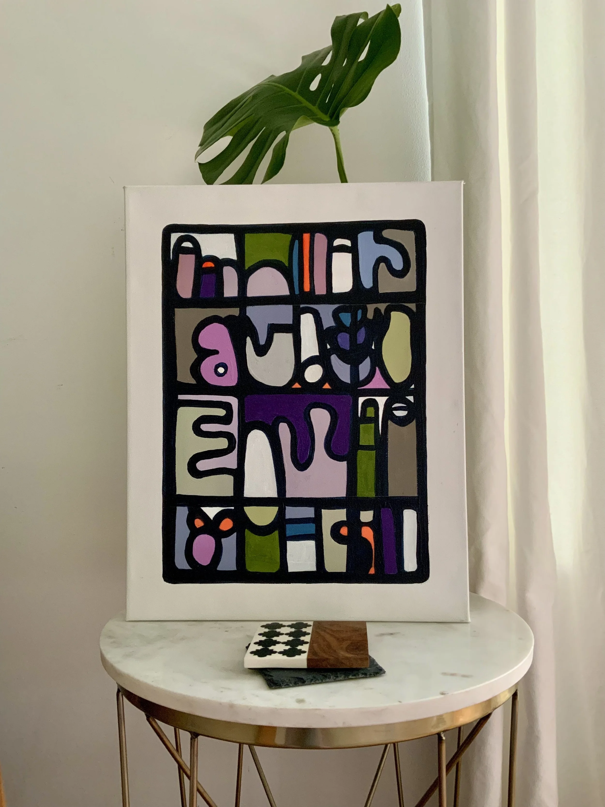 A colorful framed abstract art piece on a white wall. A green Monstera leaf is behind it. In front, a white marble-top table with a black metal frame holds a wood and ceramic chessboard with black and white squares. Denise Mortensen is a New Jersey a