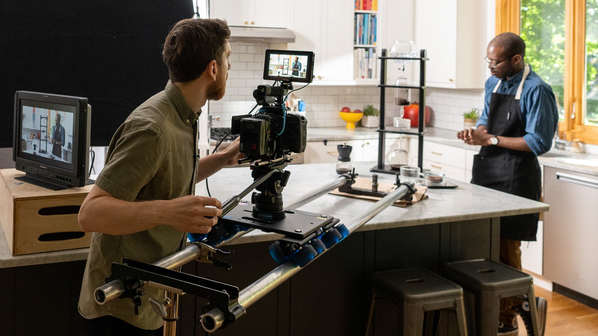 A man operates a camera on a stabilizer rig filming a man in an apron in a kitchen studio.