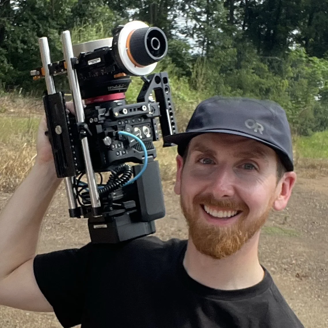 A smiling man holding a movie camera outdoors with trees and grass in the background.