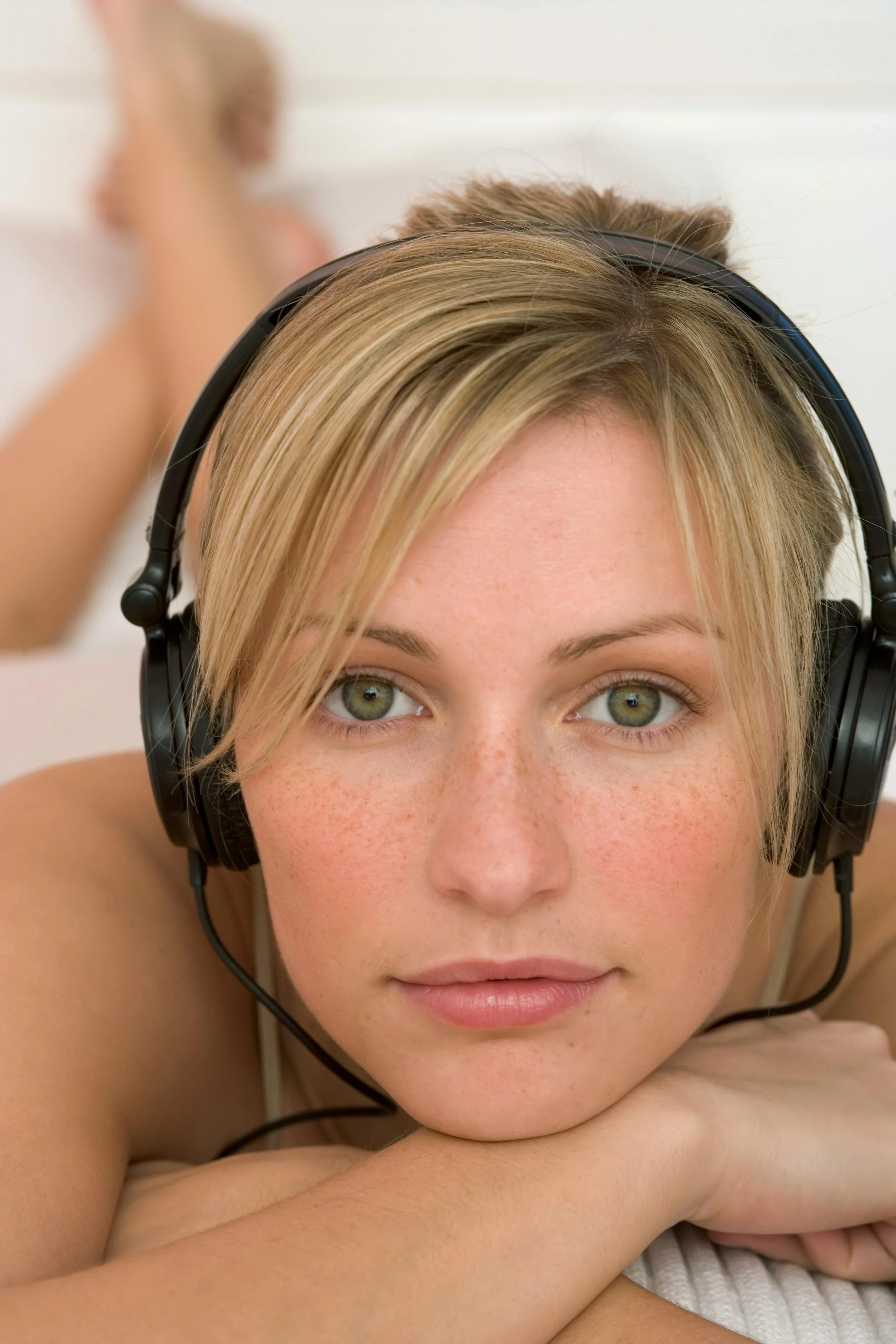 Close-up of a young woman with blonde hair, green eyes, and freckles, wearing black headphones and lying on her stomach with her chin resting on her crossed arms.