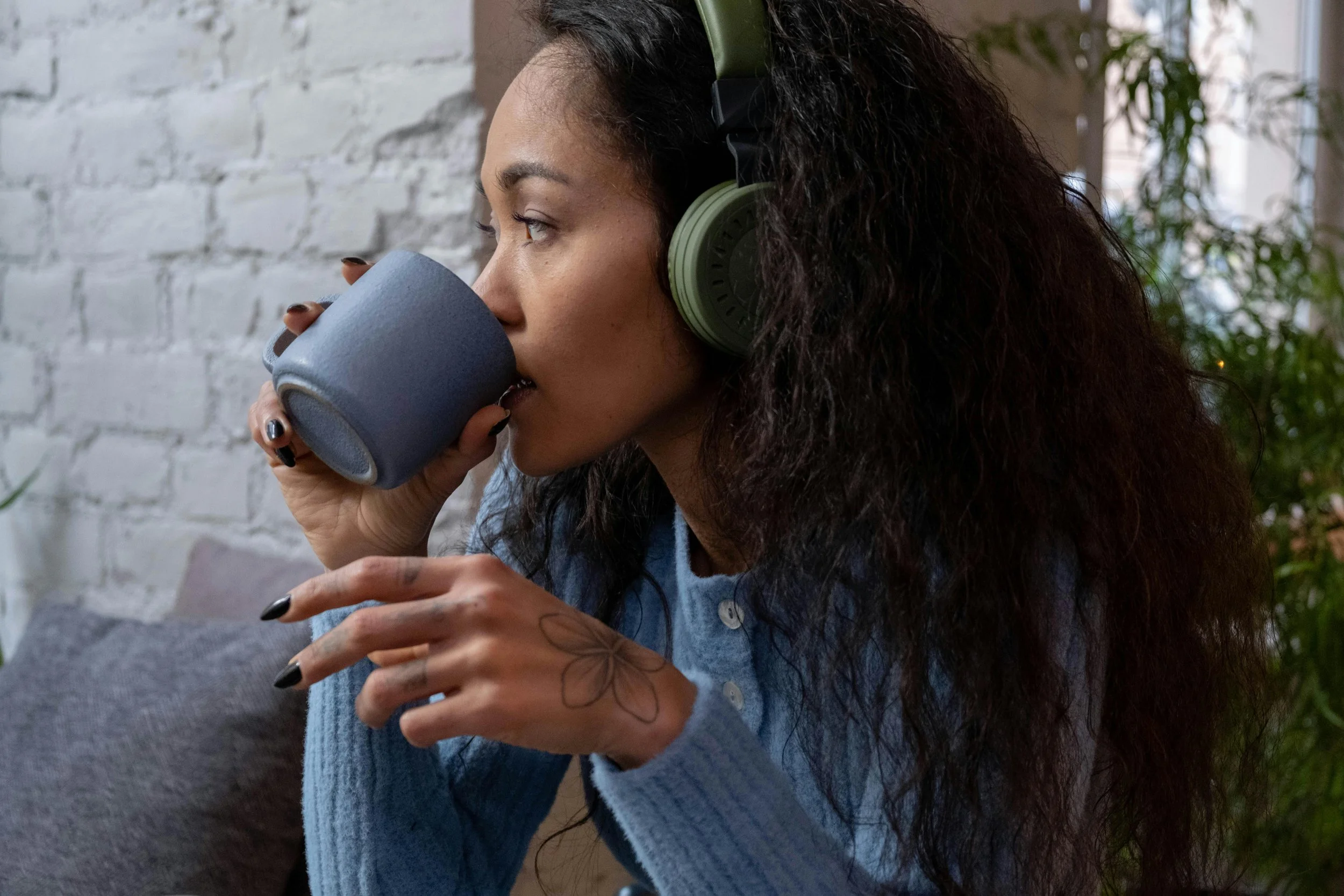 A woman with curly hair and a tattoo of a flower on her hand is sitting, drinking from a dark gray mug and wearing green over-ear headphones, with a white brick wall and green plants in the background.