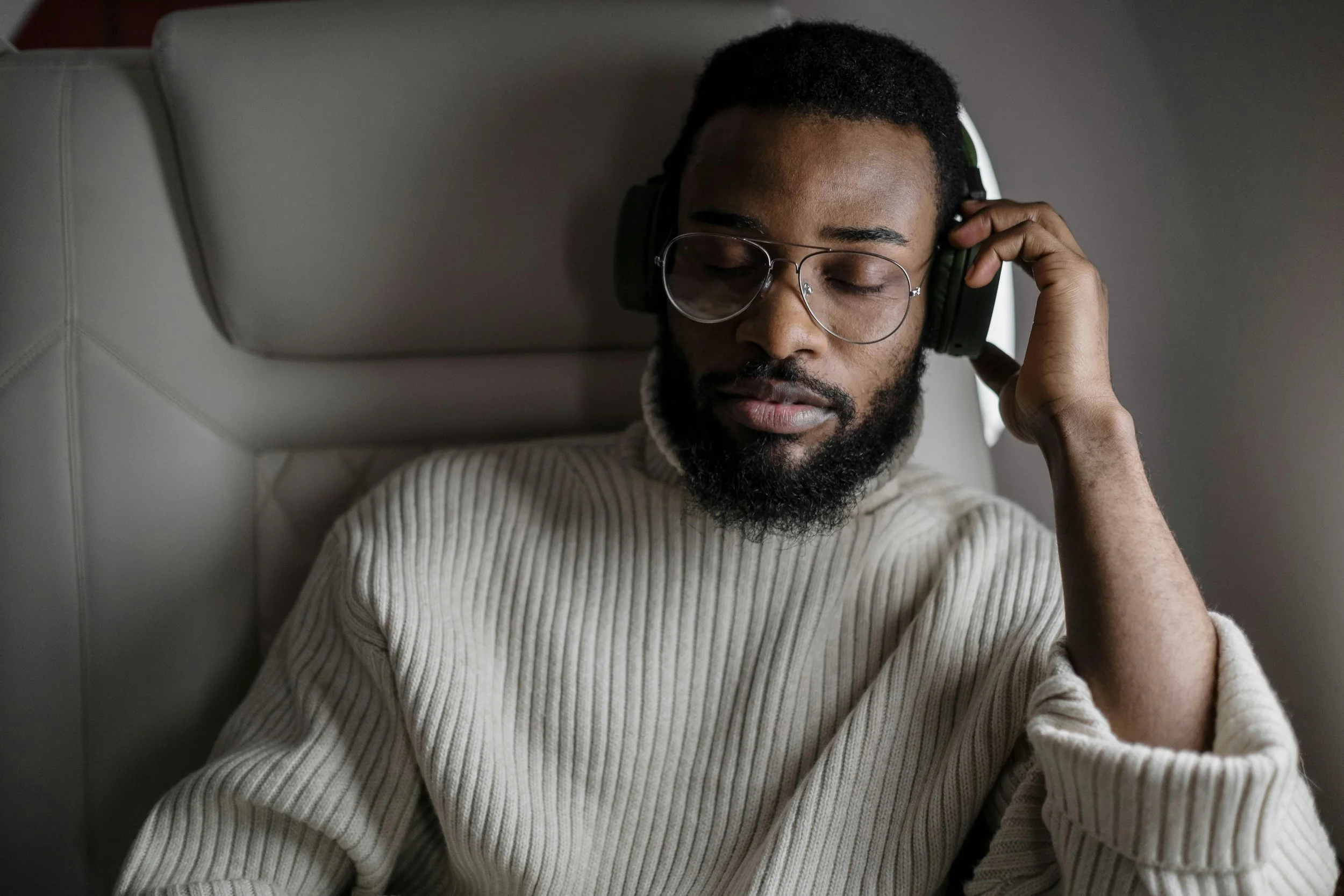 A man with glasses and a beard is wearing headphones with his eyes closed and resting against a cushioned headrest.