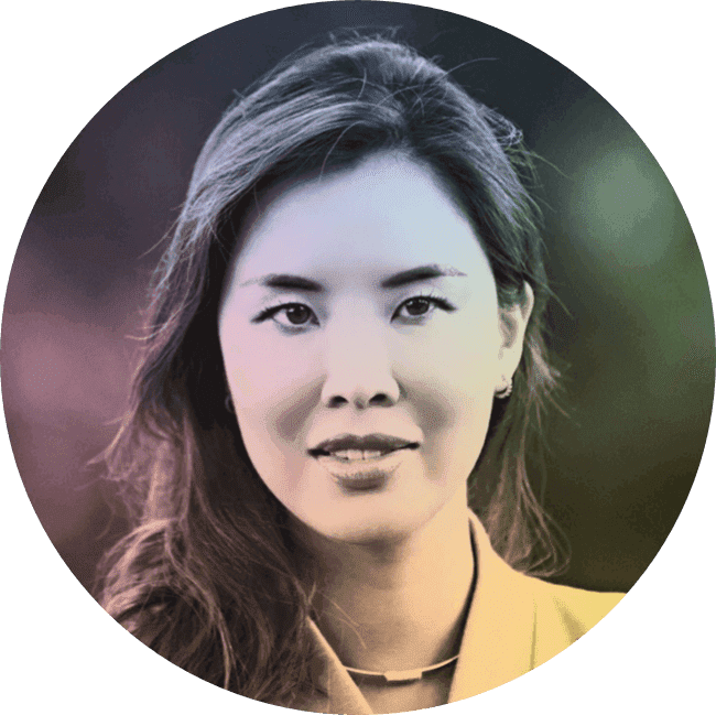 Close-up portrait of a young woman with wavy hair, wearing a light-colored jacket, posing outdoors with a blurred green background.