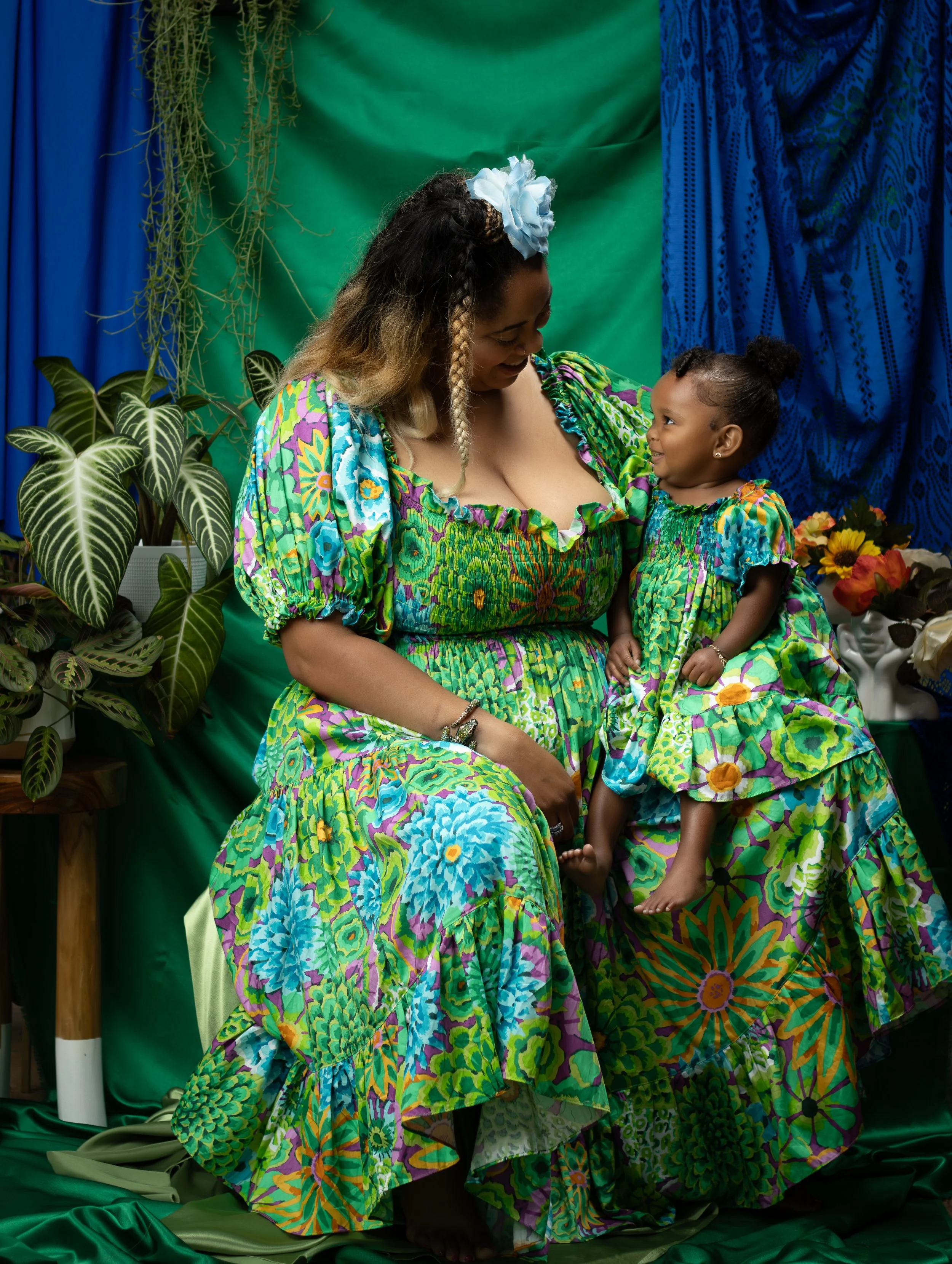 A woman and a young girl sit together, both wearing matching colorful floral dresses, surrounded by vibrant green and blue fabrics and lush plants, creating a lively and coordinated scene.