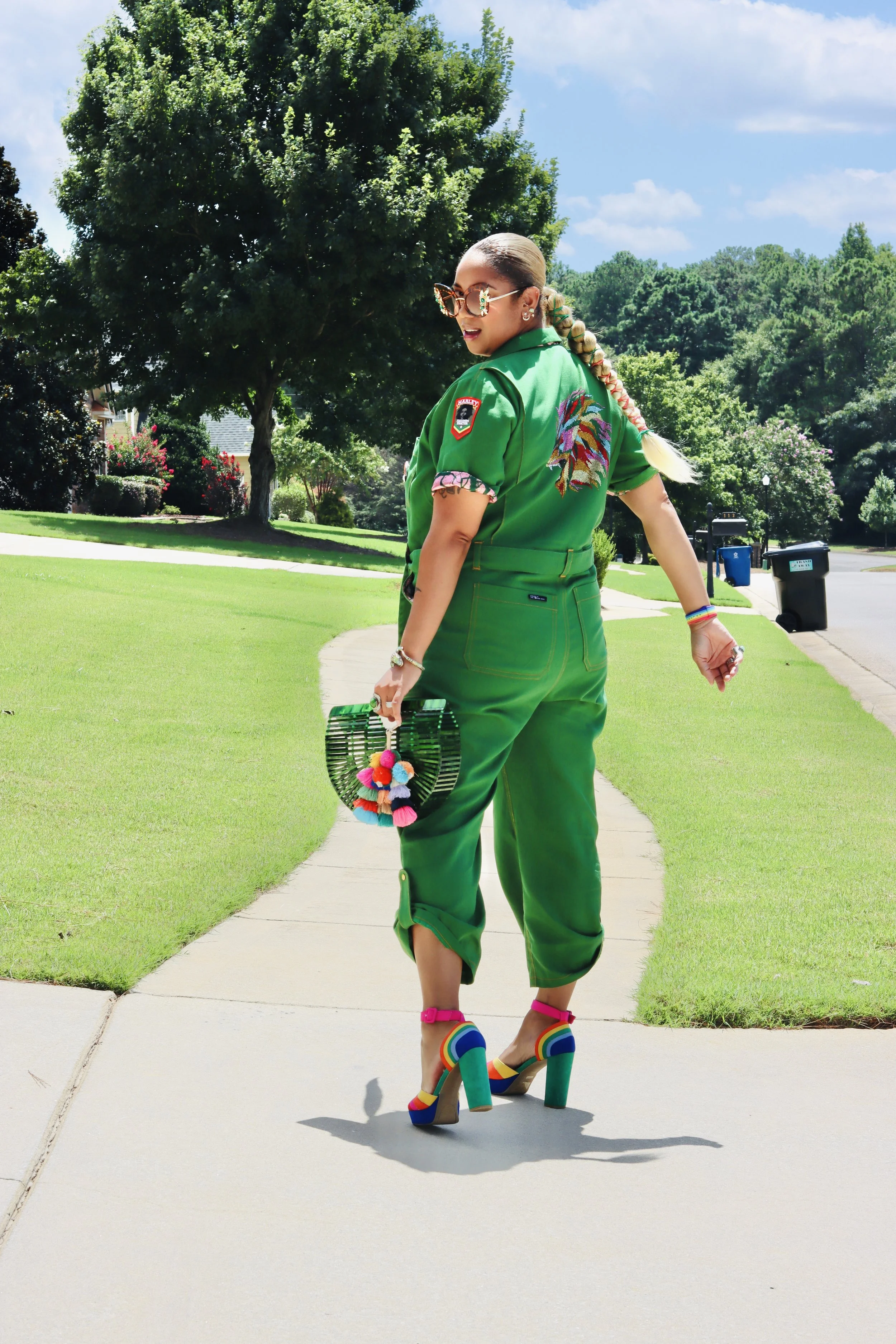 A woman walking on a sidewalk wearing a green uniform with colorful embroidery and accessories, carrying a woven purse with pom-poms, and wearing rainbow-colored high heels.