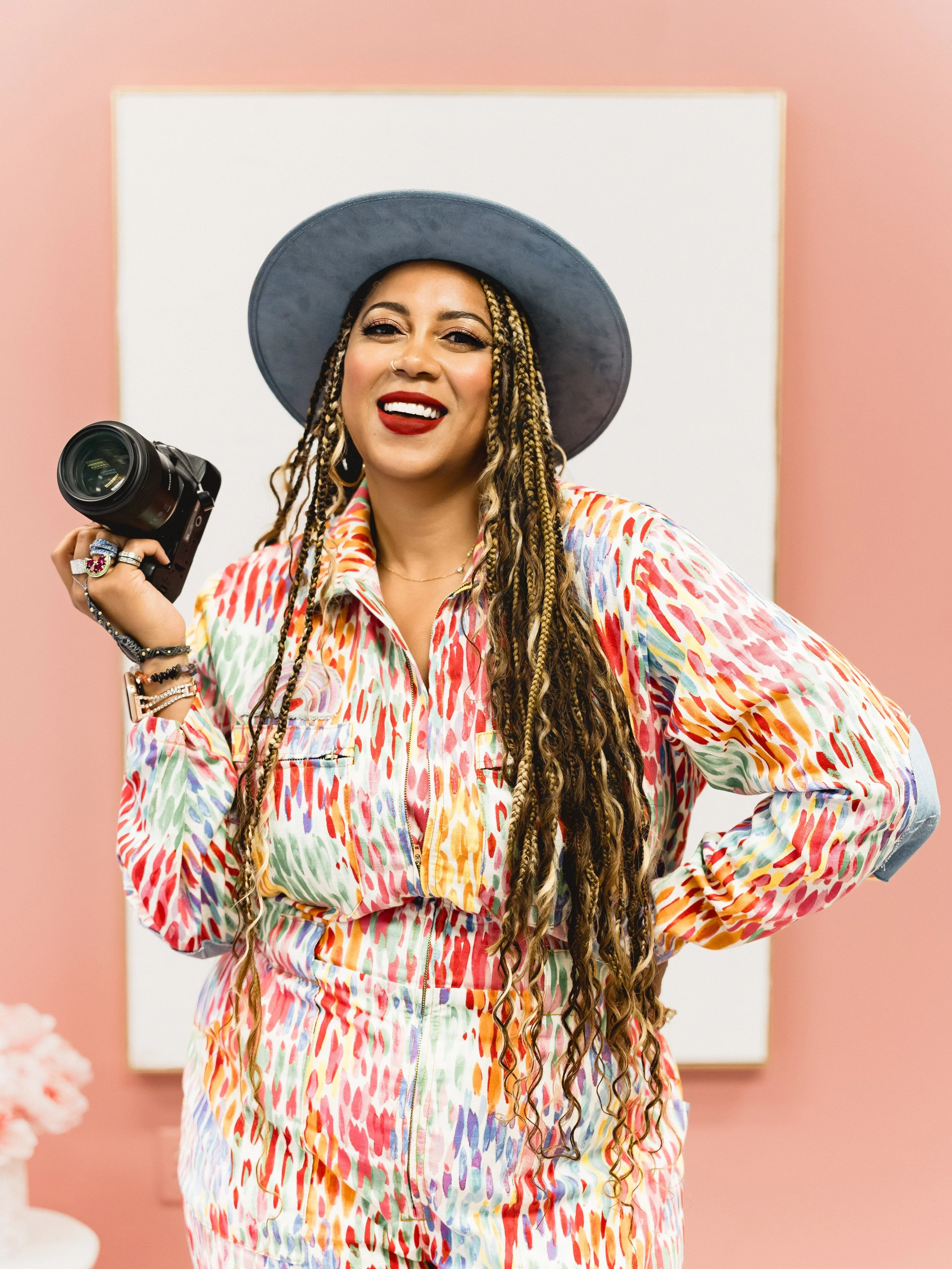 A woman with long braids, wearing a colorful patterned outfit and a wide-brimmed hat, smiling and holding a camera, standing in front of a pink wall and white board.
