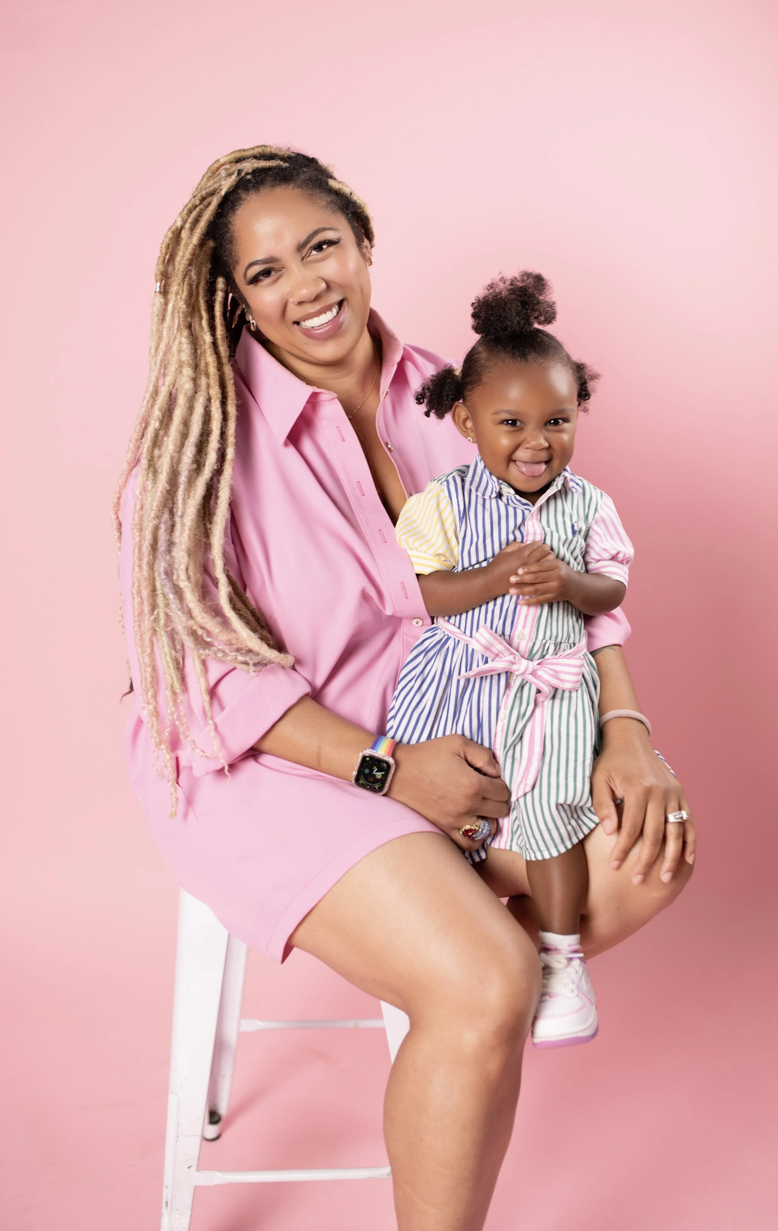 A woman with long blonde dreadlocks wearing a pink dress sitting on a white chair with a young girl on her lap, both smiling and posing for the camera against a pink background.