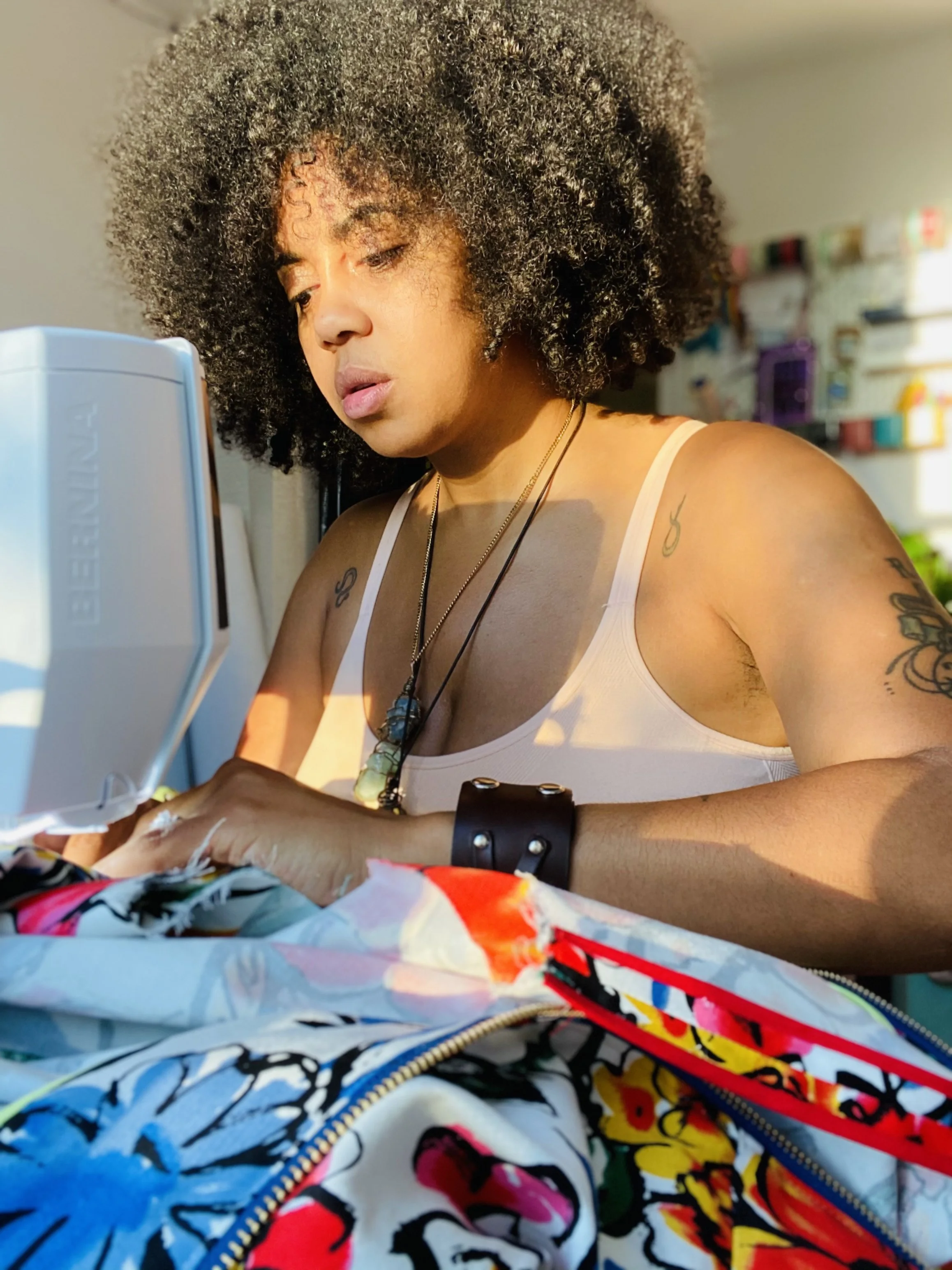 A woman with curly hair working on a sewing machine in a well-lit room, with shelves of colorful items in the background.