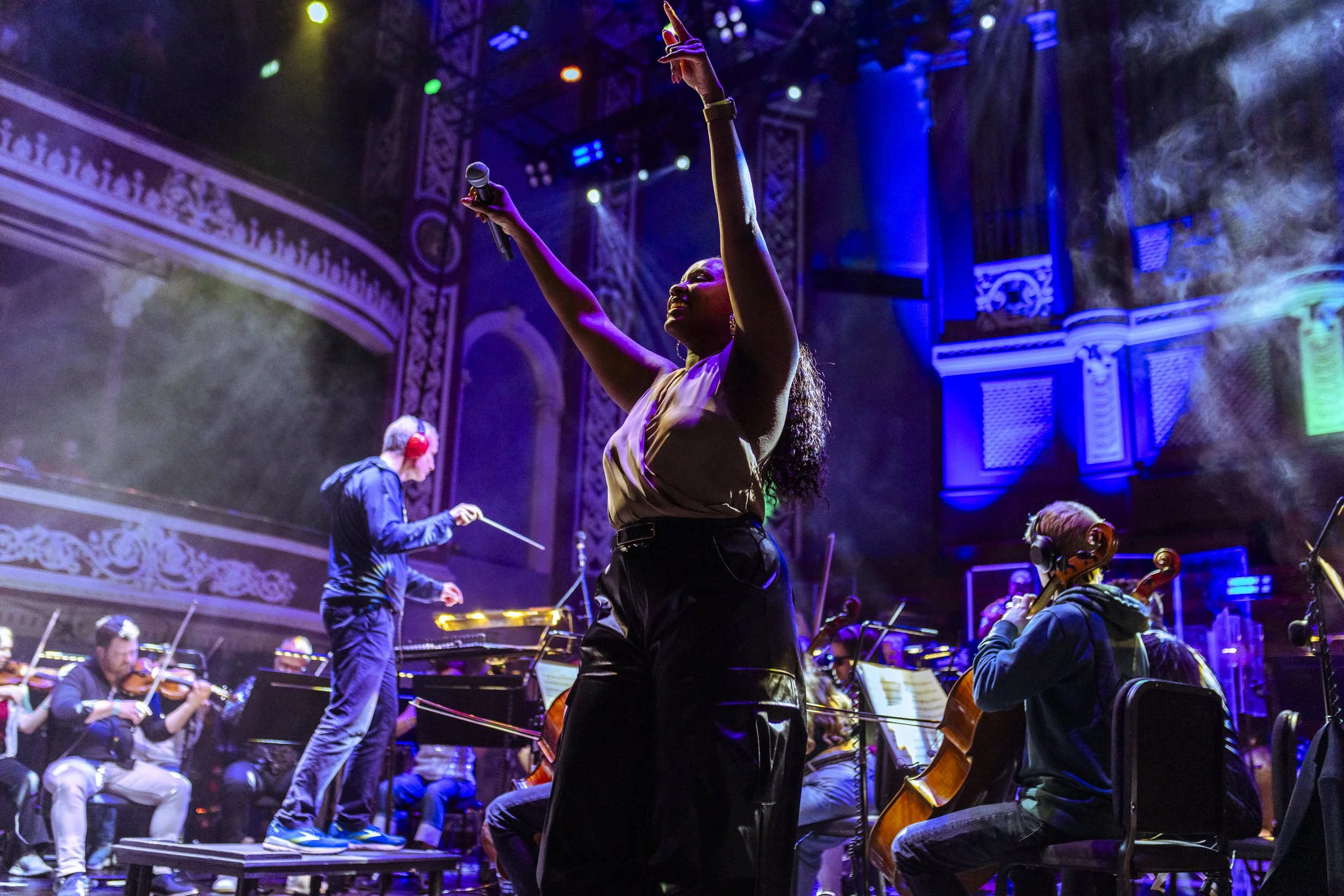 Woman singing with microphone on stage during an orchestra performance, with musicians playing violins and cellos in the background, colorful stage lighting.