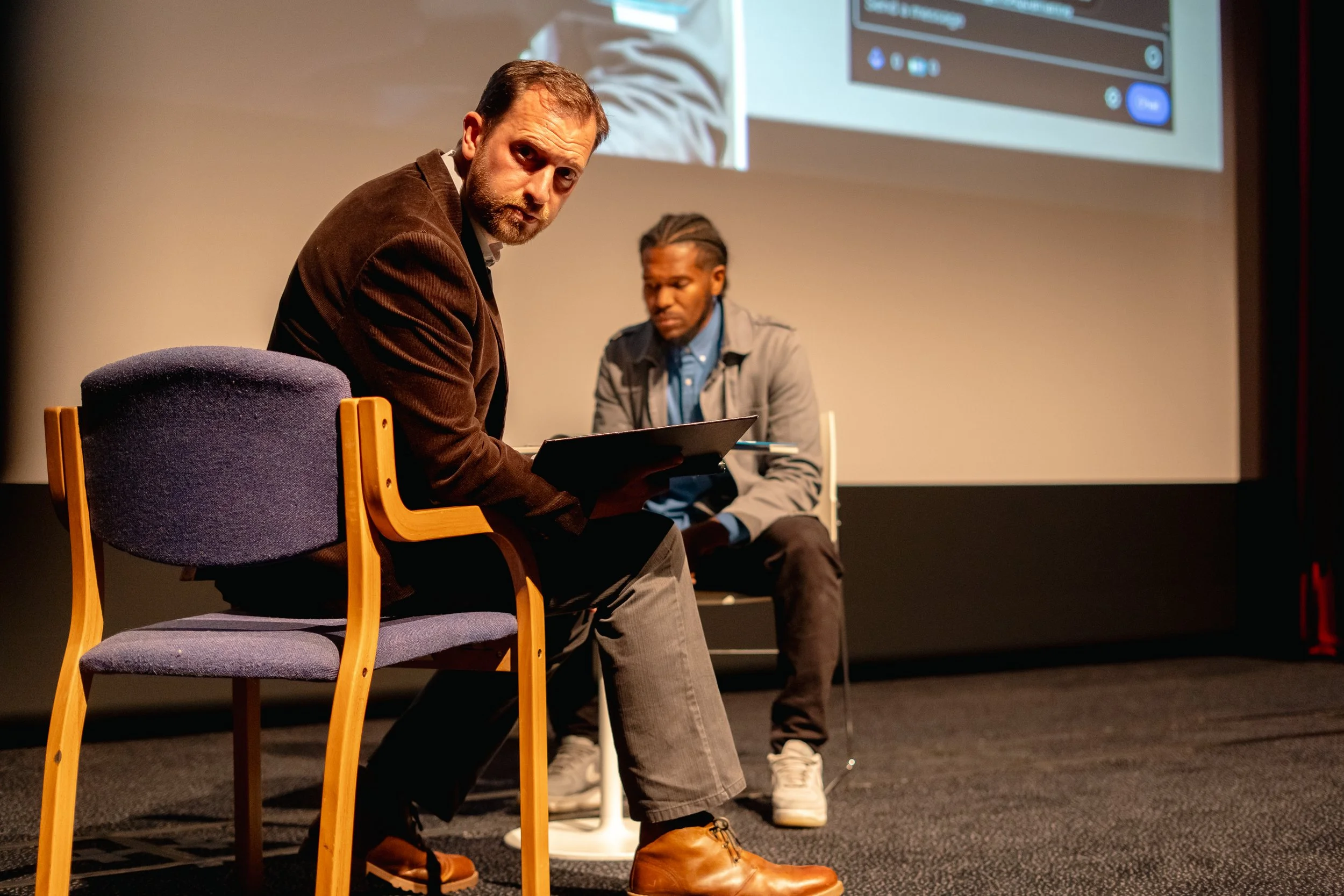 Two men sitting in chairs on stage in front of a large screen, one looking at the camera with a serious expression, the other looking down with a focused expression.