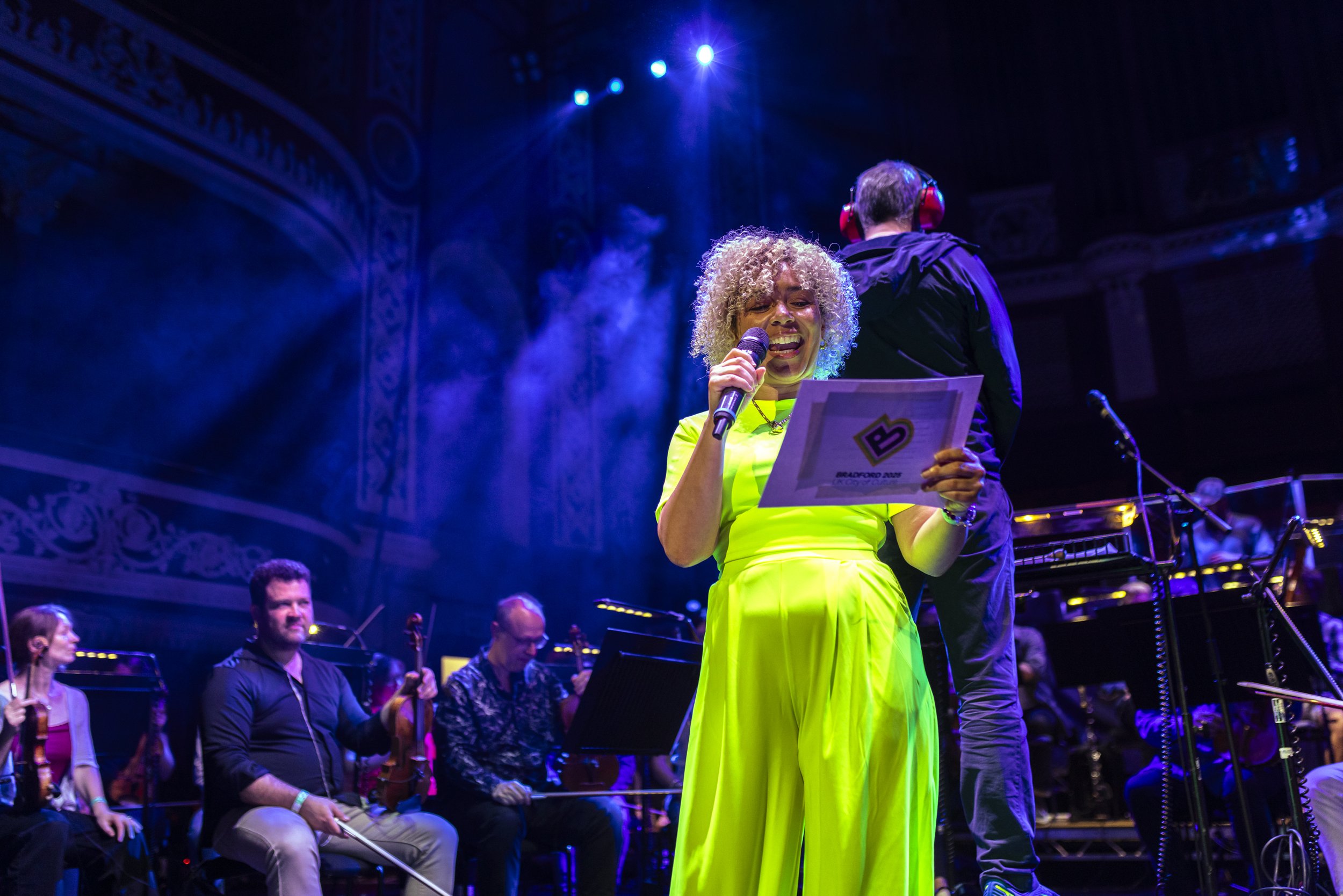 Tanya Vital stands with curly blonde hair in a bright yellow dress singing into a microphone on stage during a concert, with musicians playing string instruments seated behind her and stage lights overhead.