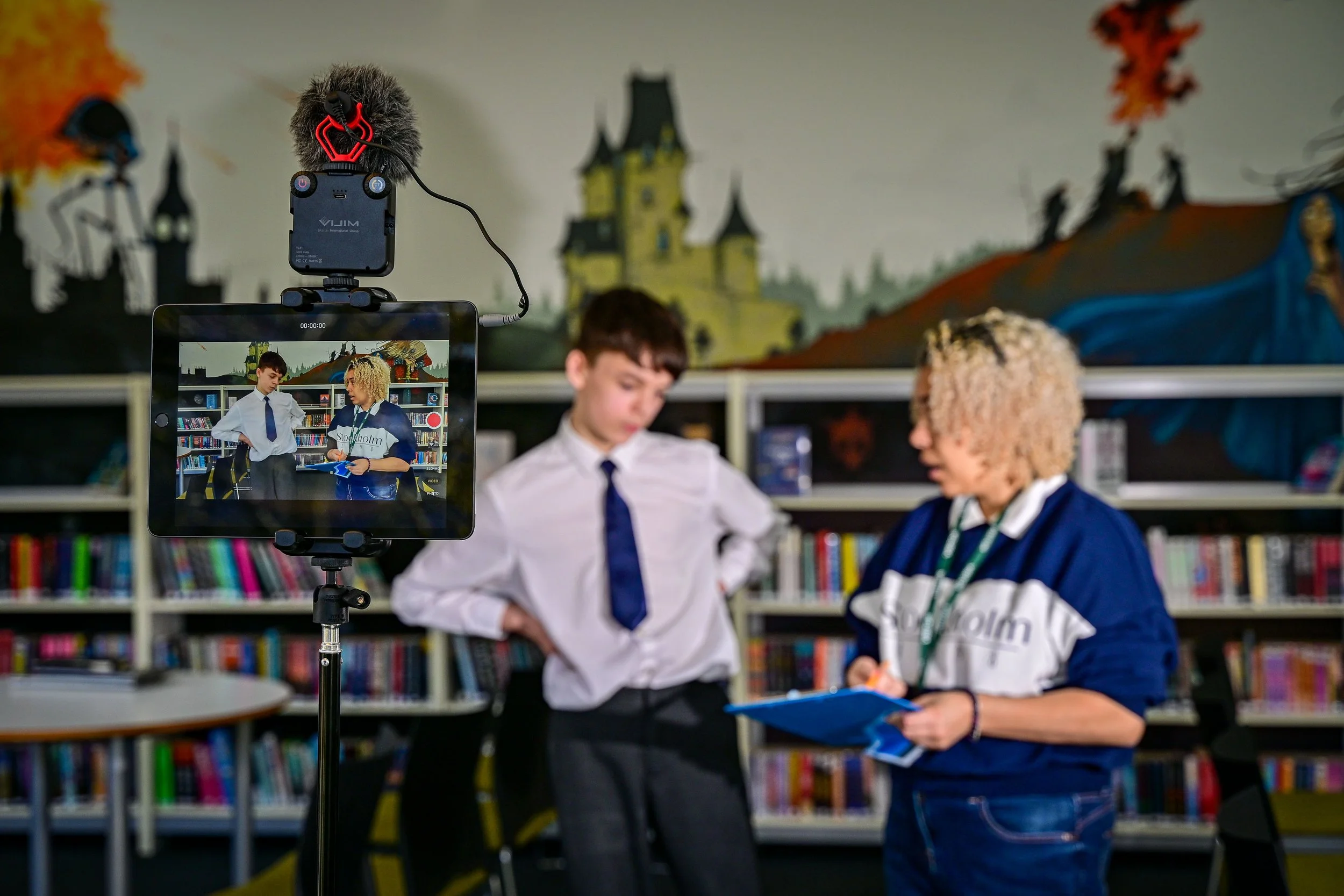 A student and a teacher in a library recording a video with a tablet mounted on a tripod. The tablet screen shows the same scene, with bookshelves and a colorful mural of a haunted house in the background.