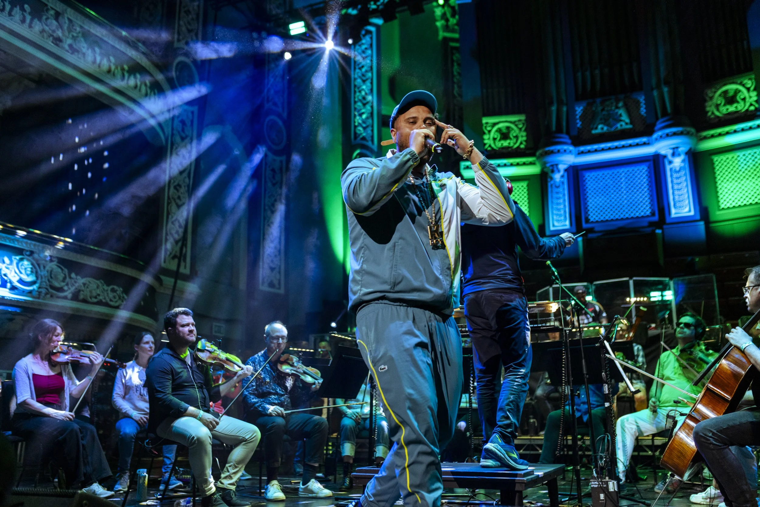 K Dot in a tracksuit and cap singing into a microphone on a stage with an orchestra behind him in a concert hall with ornate green and blue decorations.