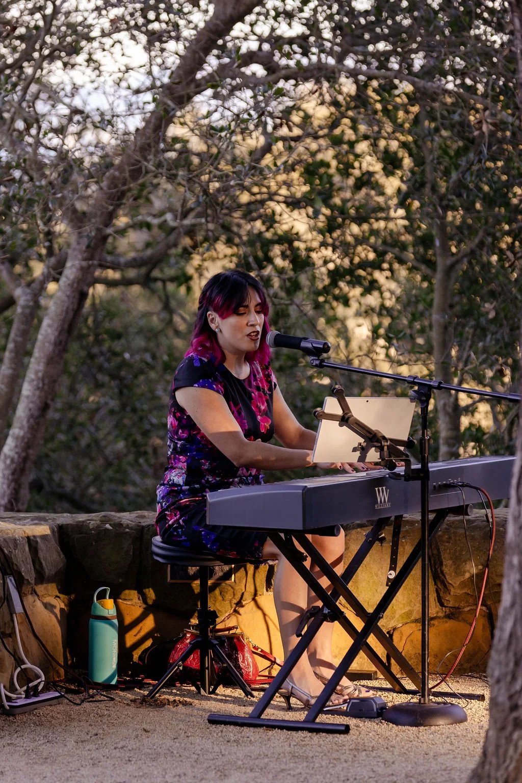 A woman with dark and pink hair singing and playing a keyboard outdoors in a wooded area during sunset.