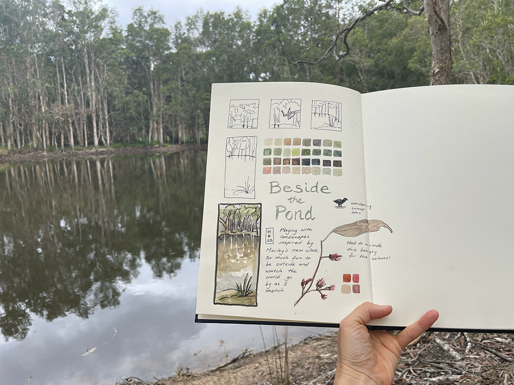 A sketchbook with a landscape  painting of a pond, held up in front of the pond that has been painted.