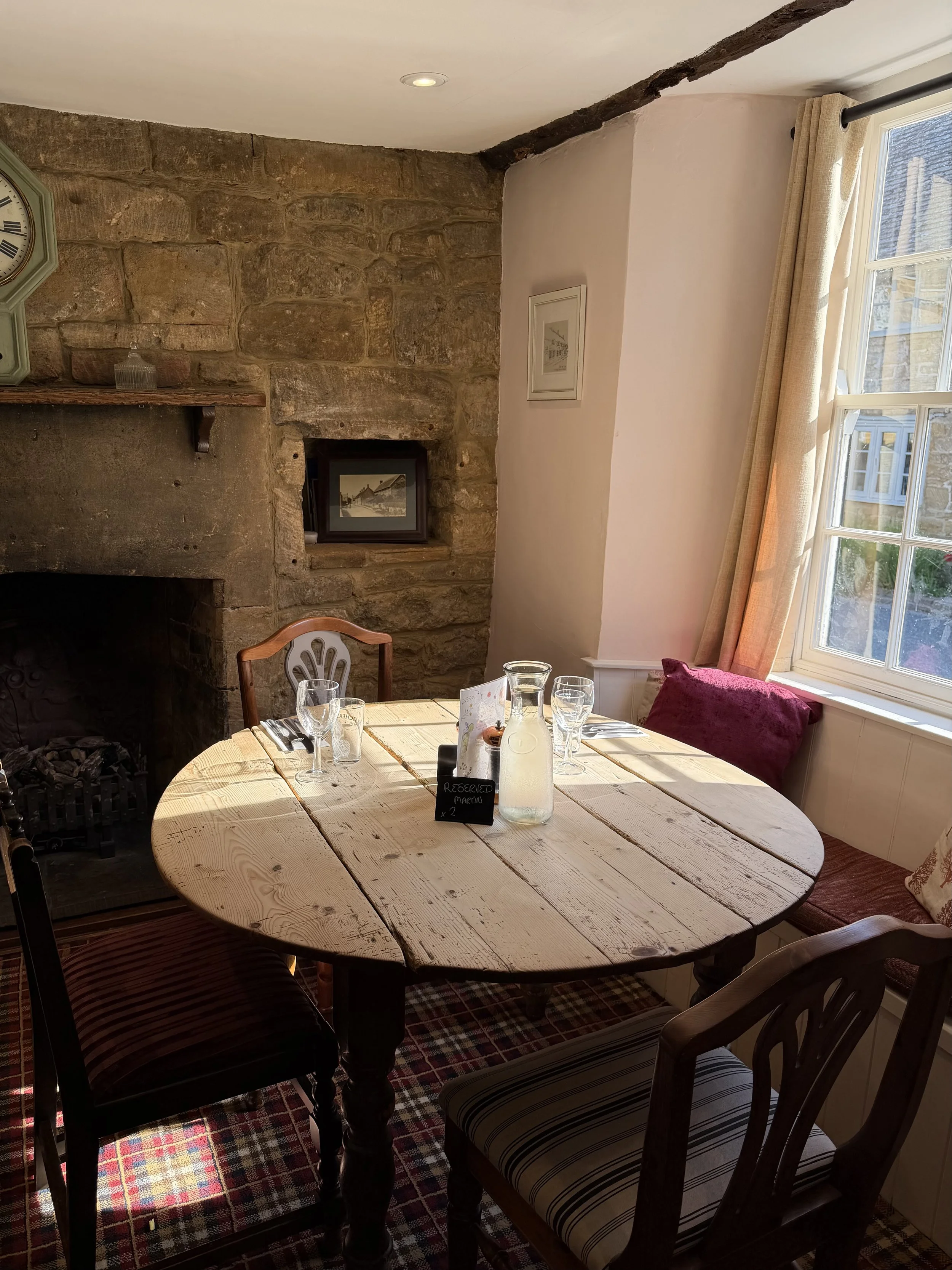 A cozy dining room with a round wooden table set with glasses and a bottle of water, surrounded by chairs with striped cushions, a stone fireplace with a wooden mantel, framed pictures on white walls, large windows with beige curtains, and a checkered rug on the floor.
