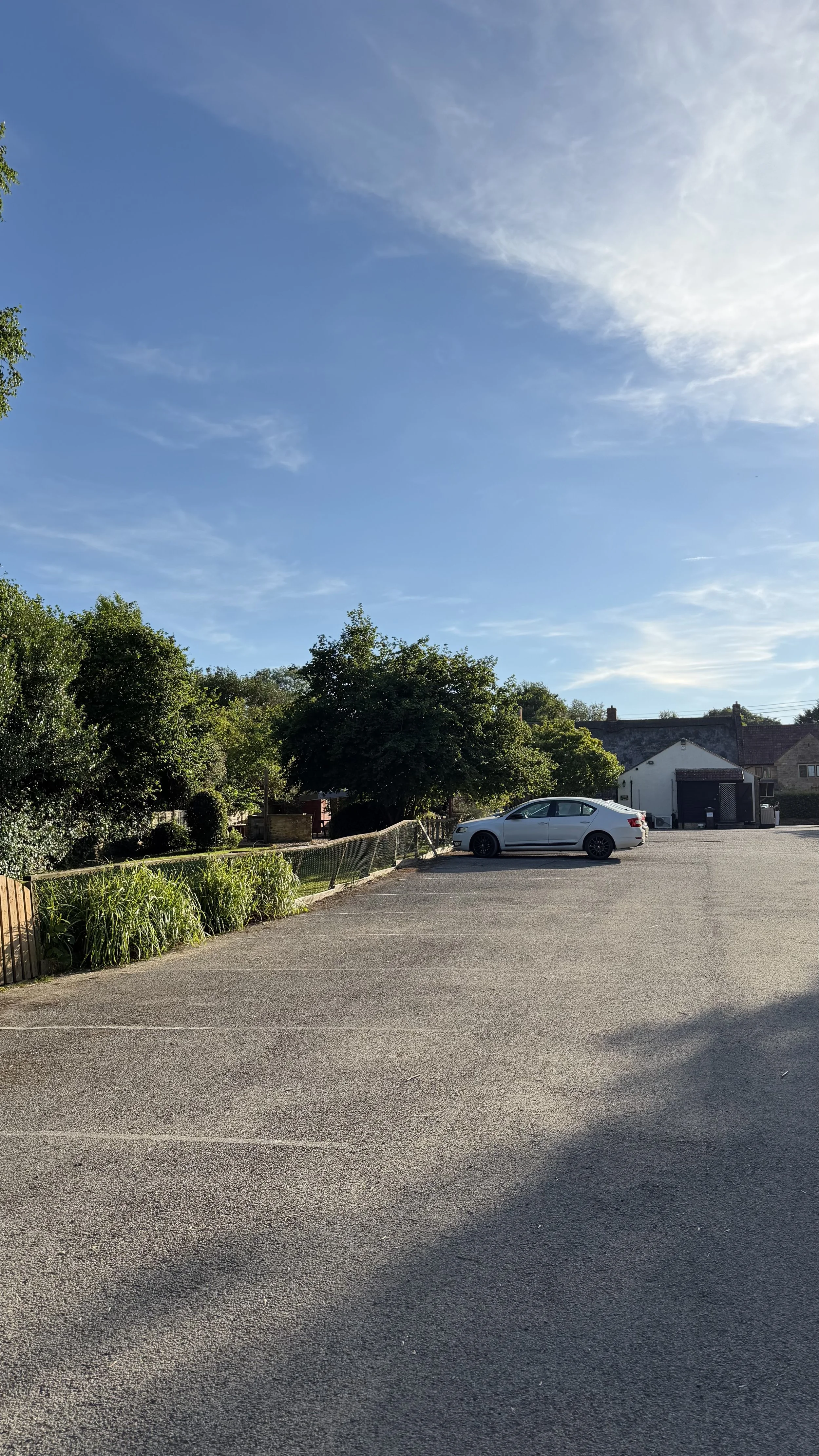 A parking lot with a single white car parked next to trees and bushes. Behind the trees are houses and a blue sky with some clouds.