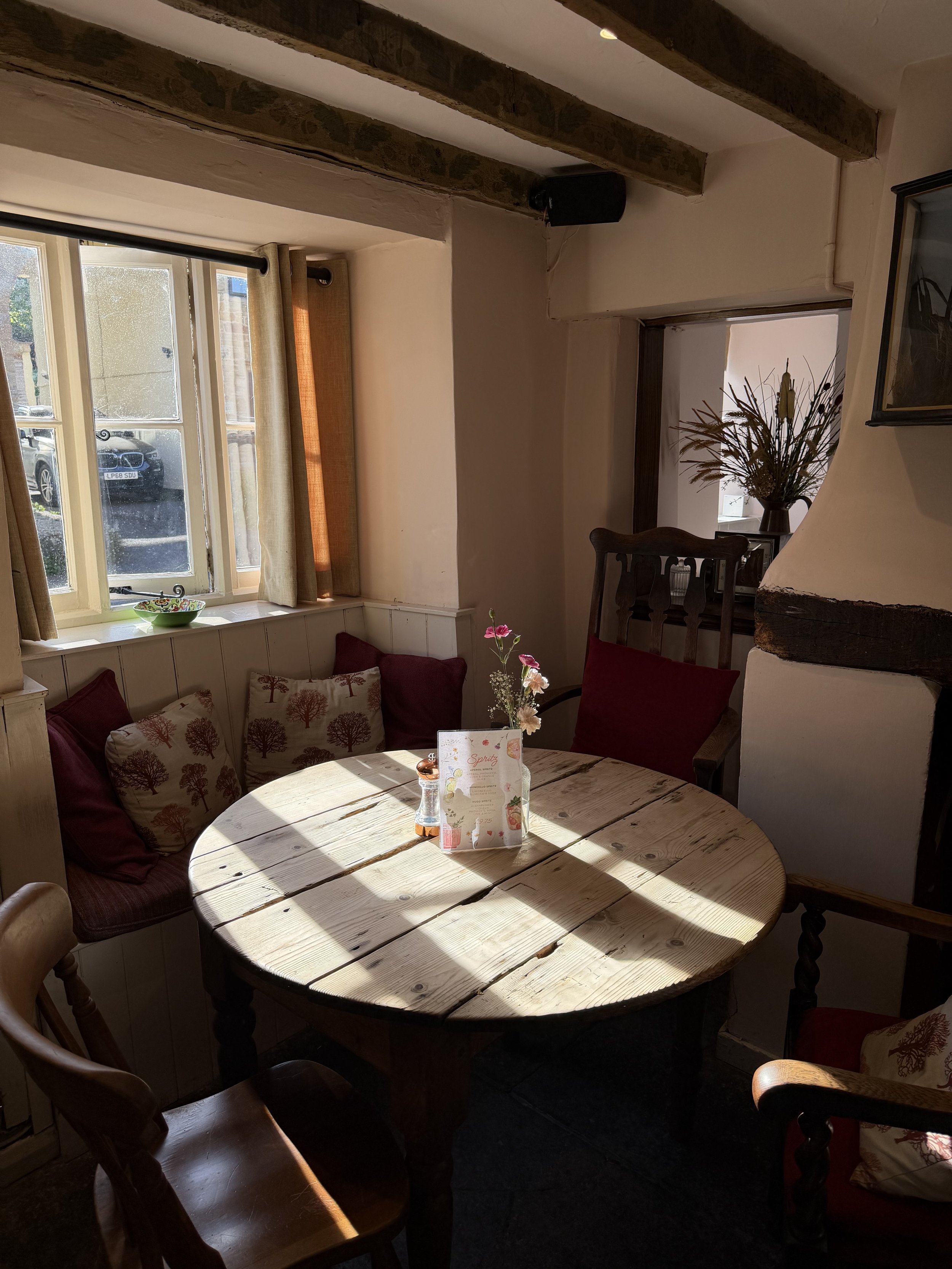A dining area in a cozy restaurant with sunlight coming through the window, a round wooden table with a vase of pink flowers, and cushioned chairs around it, decorated with pillows featuring tree motifs.