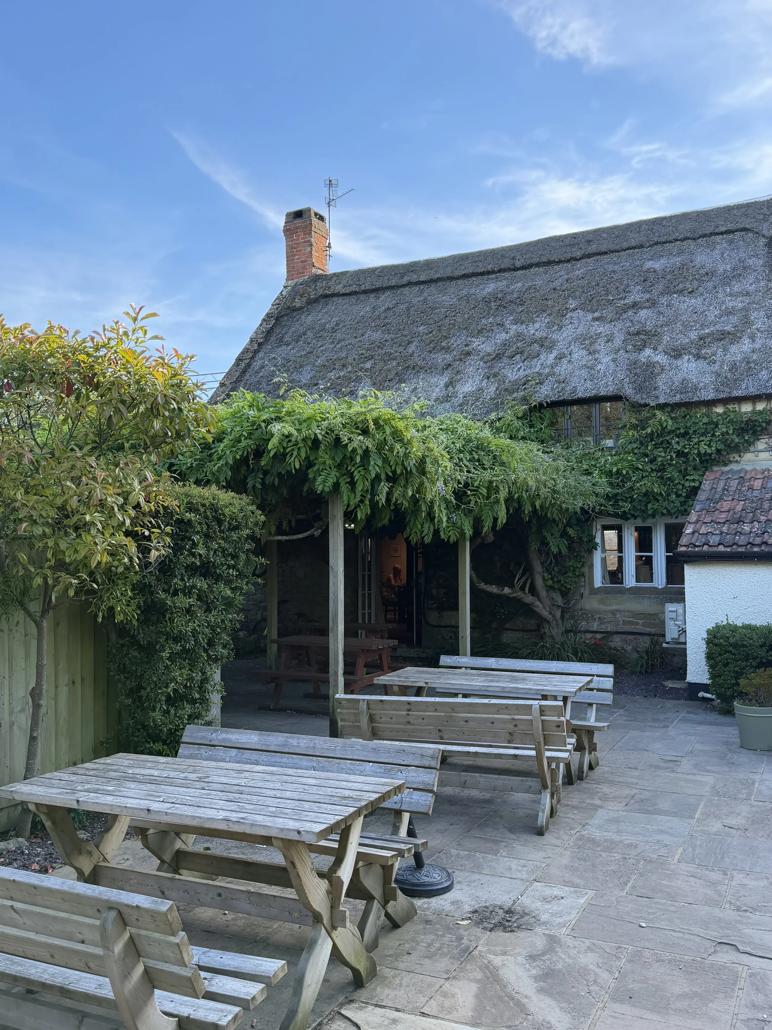 An outdoor patio area with wooden tables and benches, surrounded by greenery and a house with a thatched roof. There is a fence and a small window on the house. The sky is blue with some clouds.