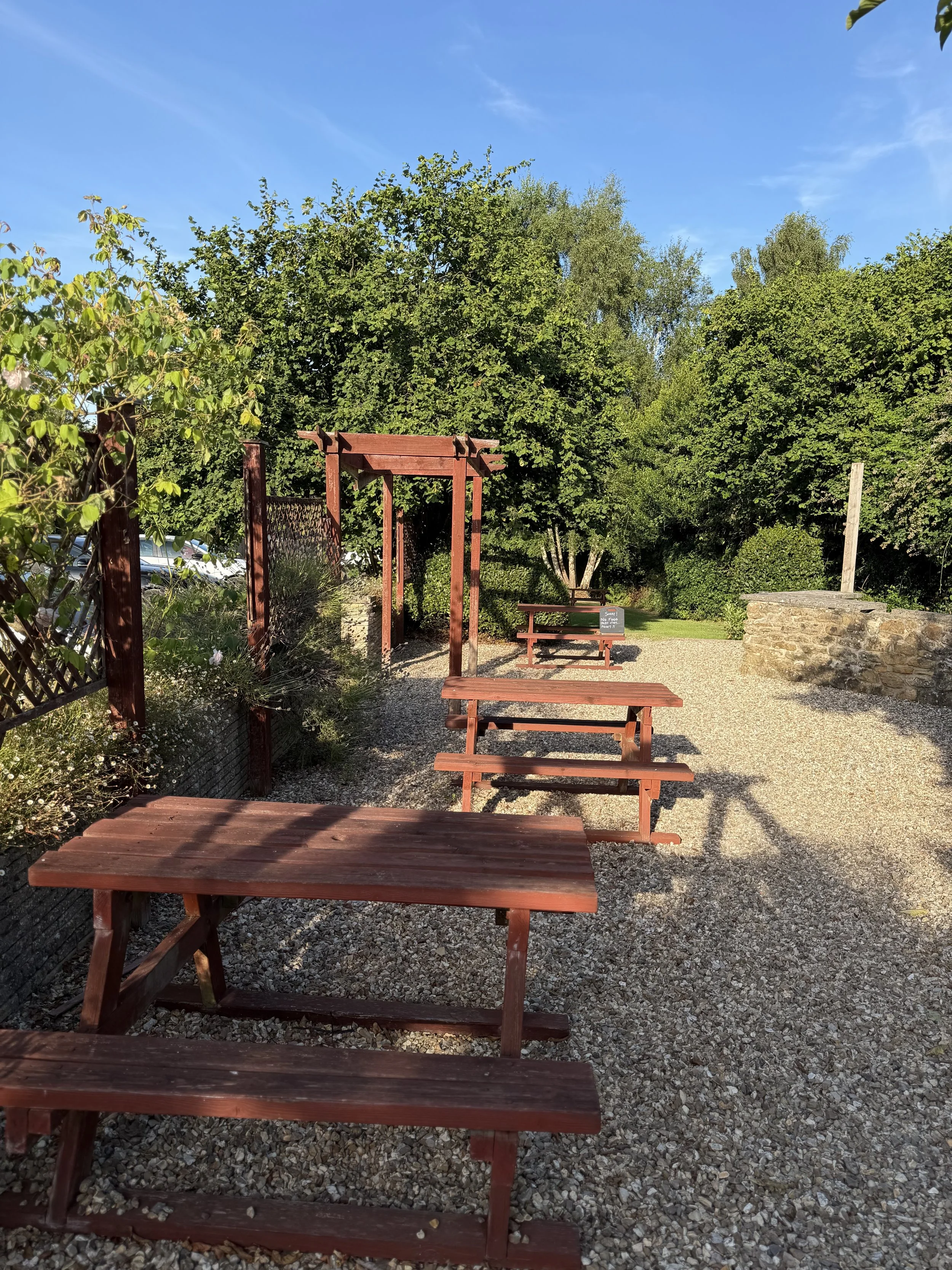 Outdoor scene with red wooden benches, a pergola, surrounded by green trees, a stone wall, and a gravel ground, under a blue sky.