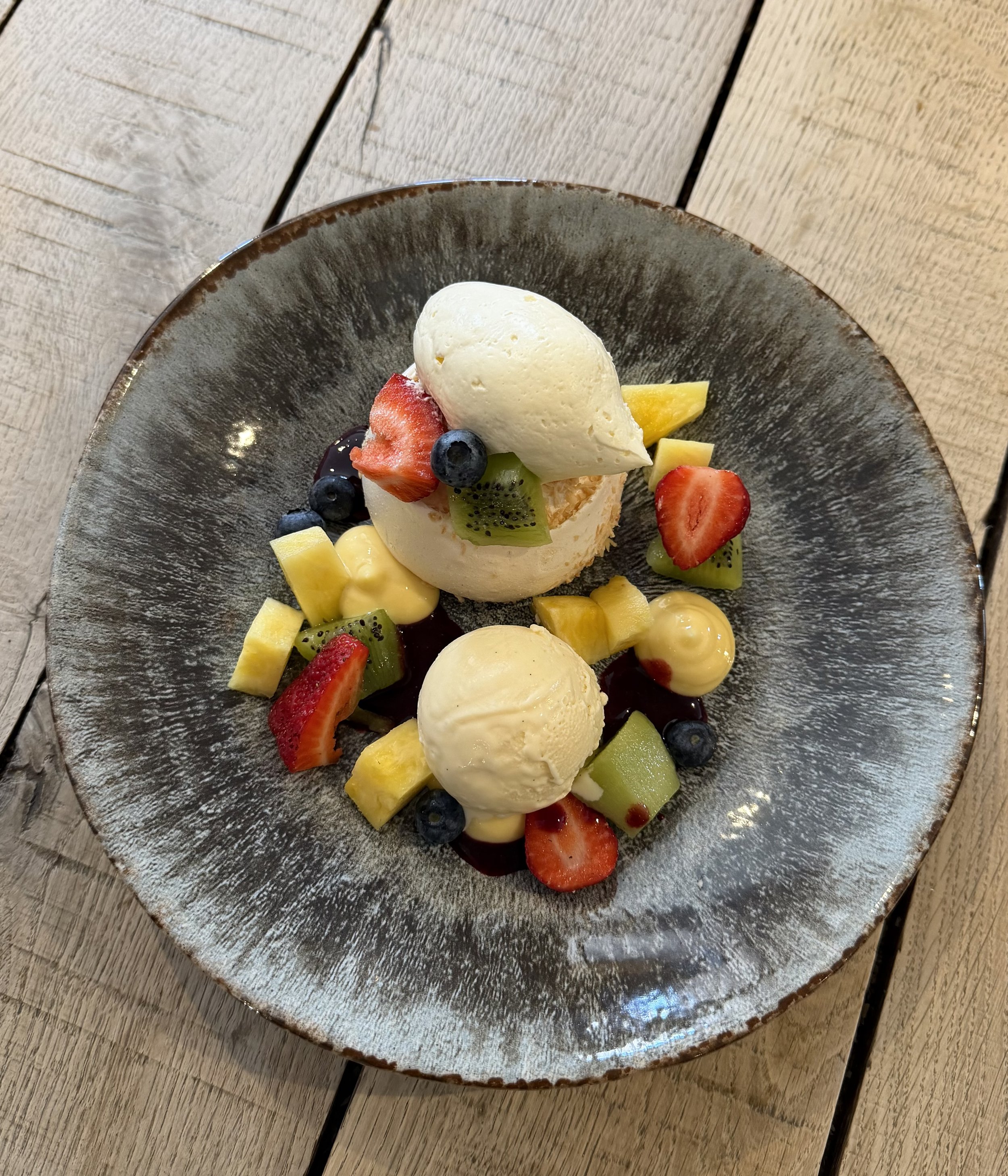 A dessert plate holding two scoops of vanilla ice cream, surrounded by sliced strawberries, kiwi, mango, and blueberries, with a piece of lemon and a drizzle of berry syrup on a rustic wooden table.