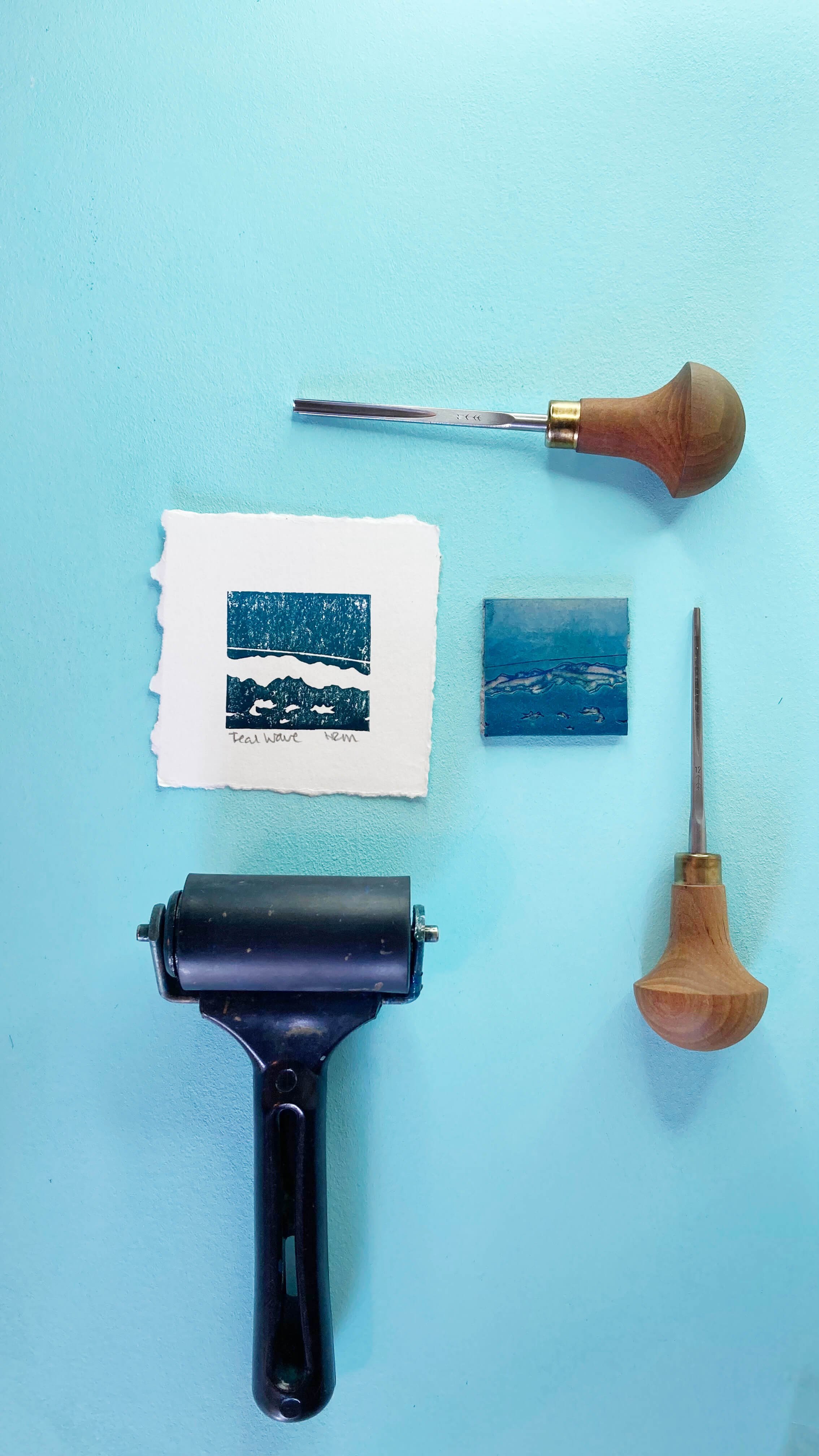 'Teal Wave' sea print next to the tiny Lino block, Lino gouges and black brayer against a blue background.