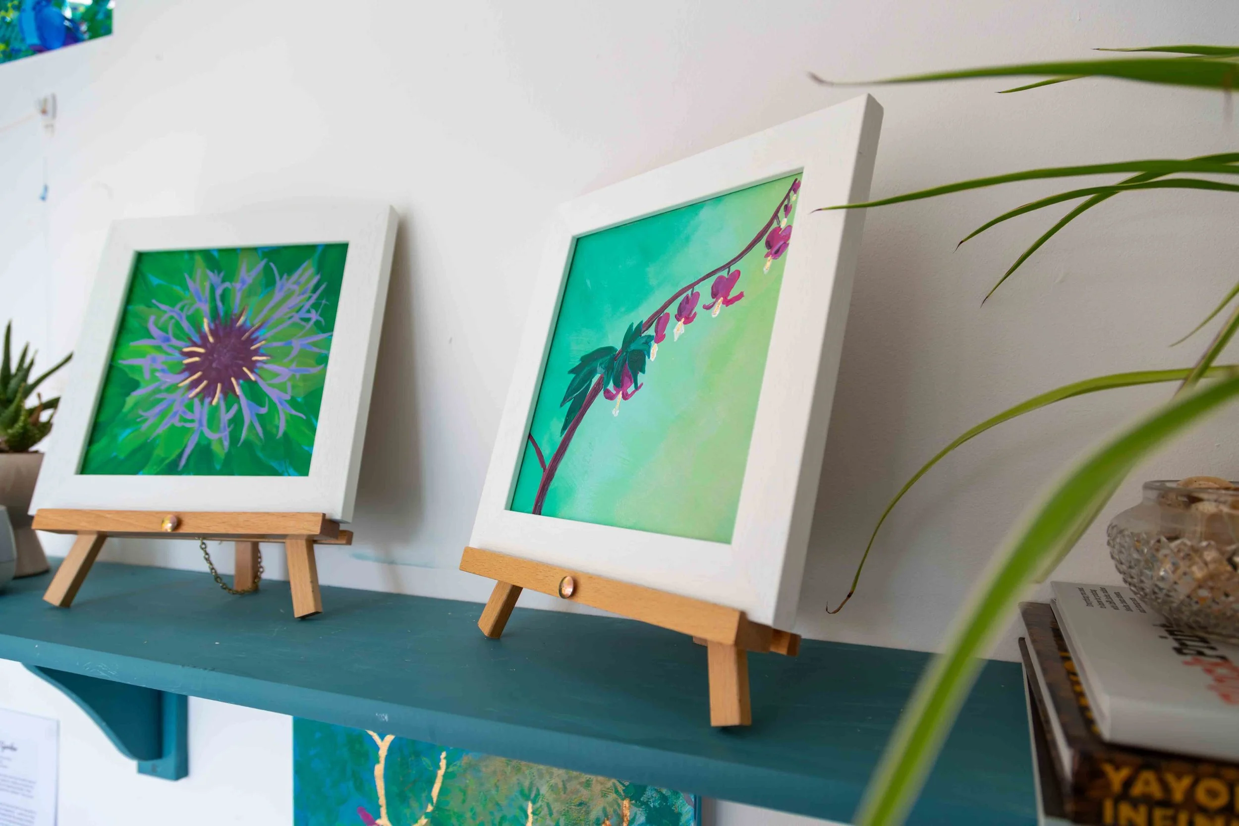 Two floral artworks in white frames on wooden easels atop a teal painted shelf on a white studio wall