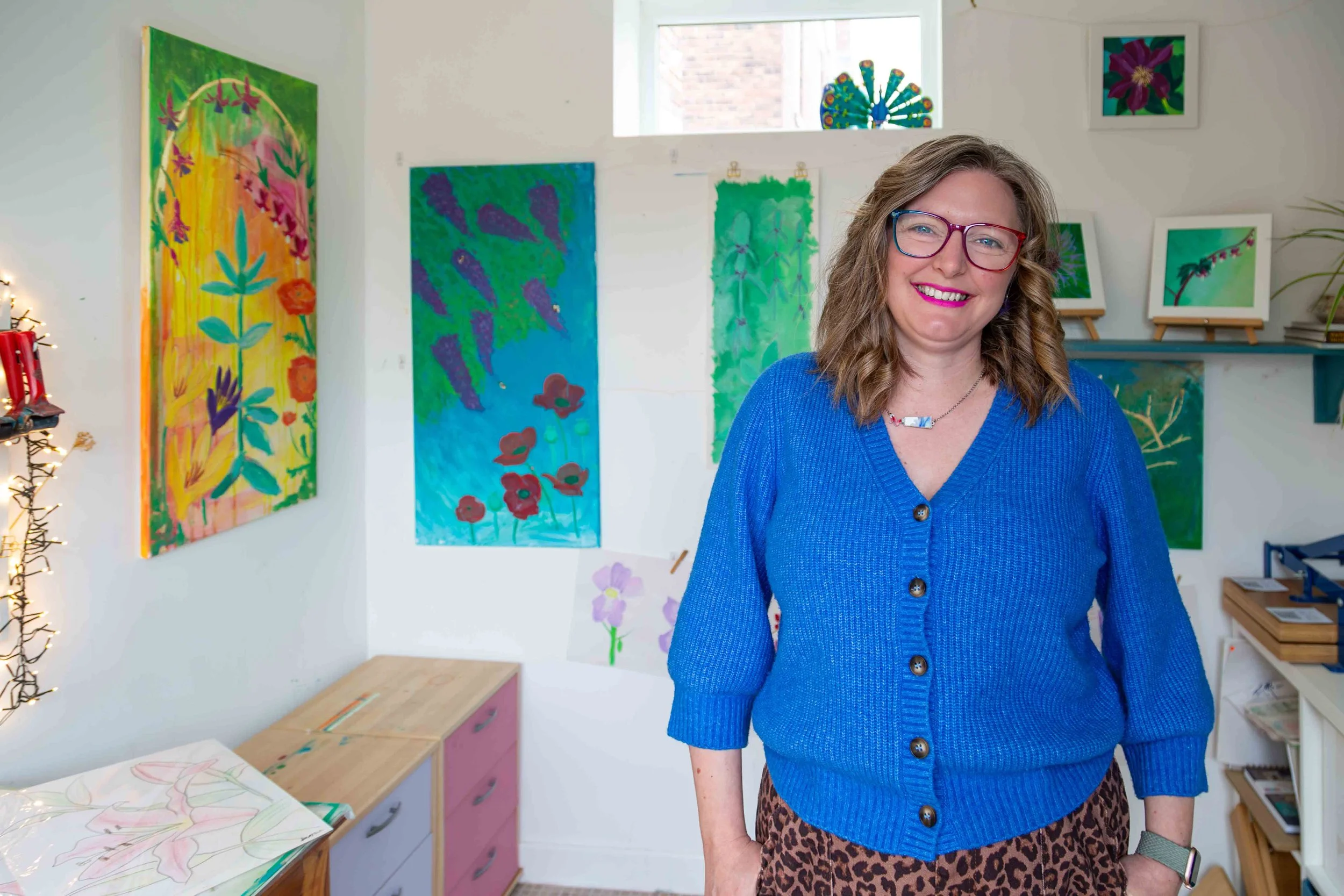 Heather Moore, a blonde woman in blue cardigan and leopard print trousers, is stood in her studio with colourful landscape artworks behind her