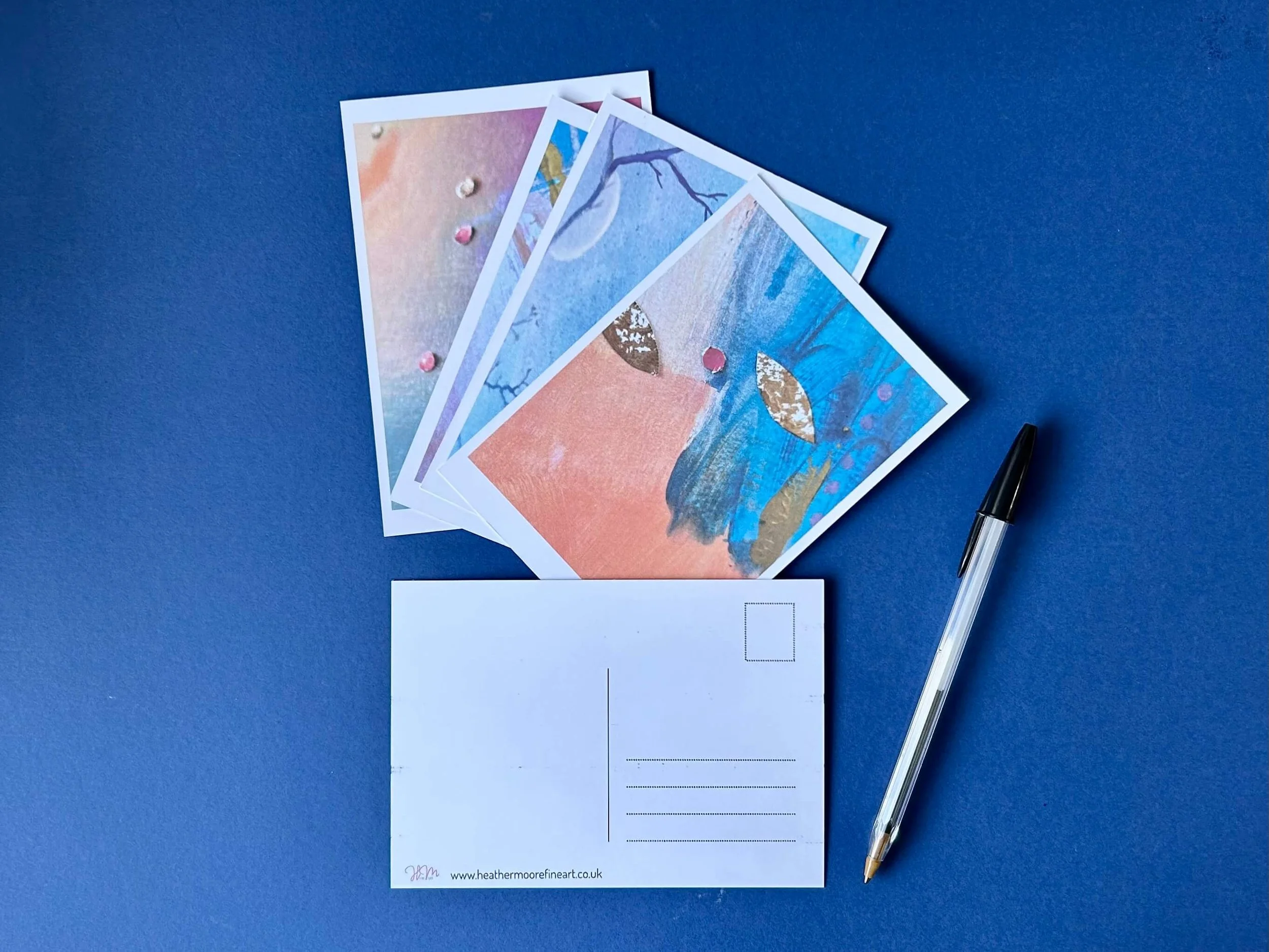 4 art postcards fanned on a blue surface and one turned over ready to write with a black biro