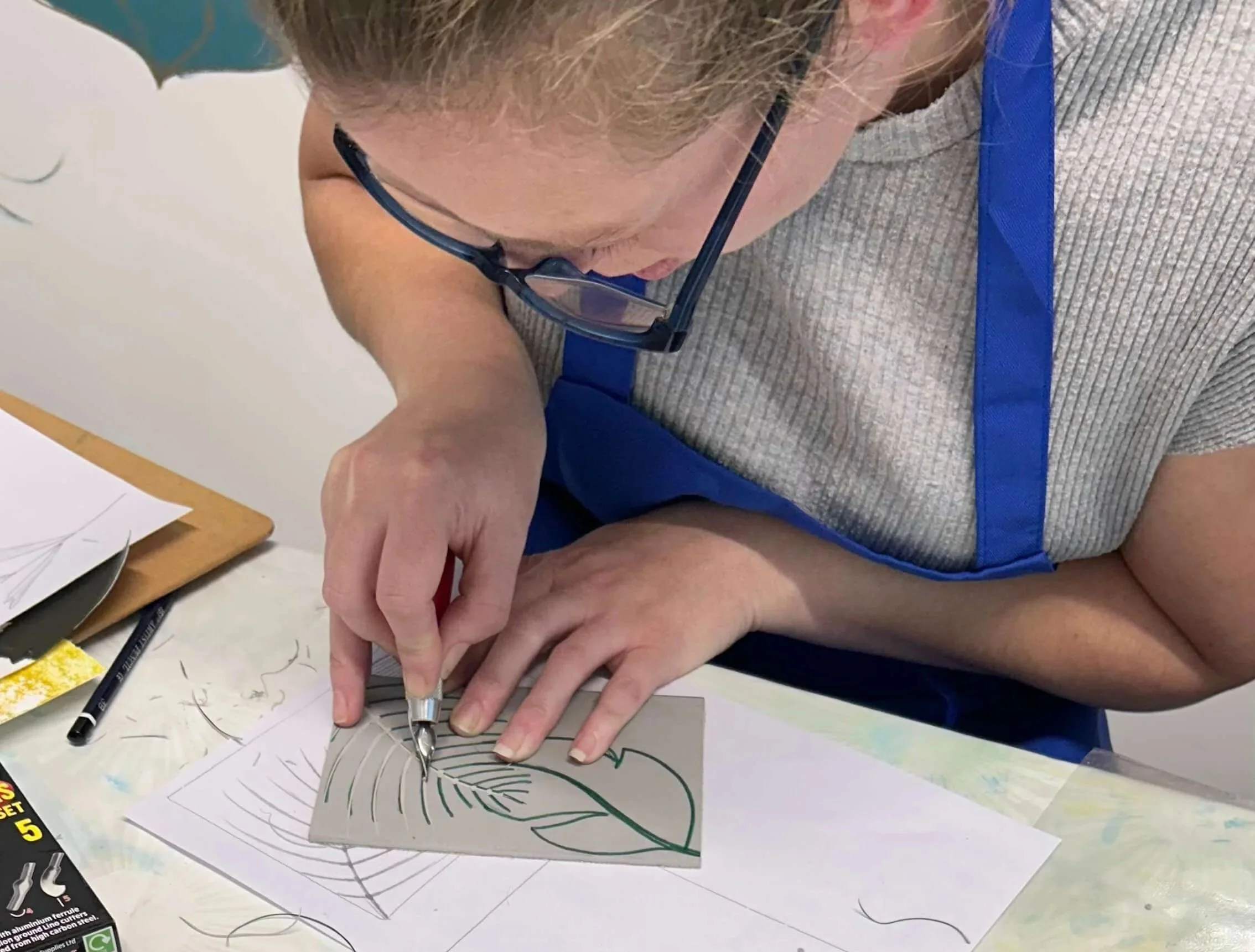 A person wearing glasses and a gray shirt is using a Lino gouge to carve a line into a Lino block with a leaf design, placed on a white sheet of paper.