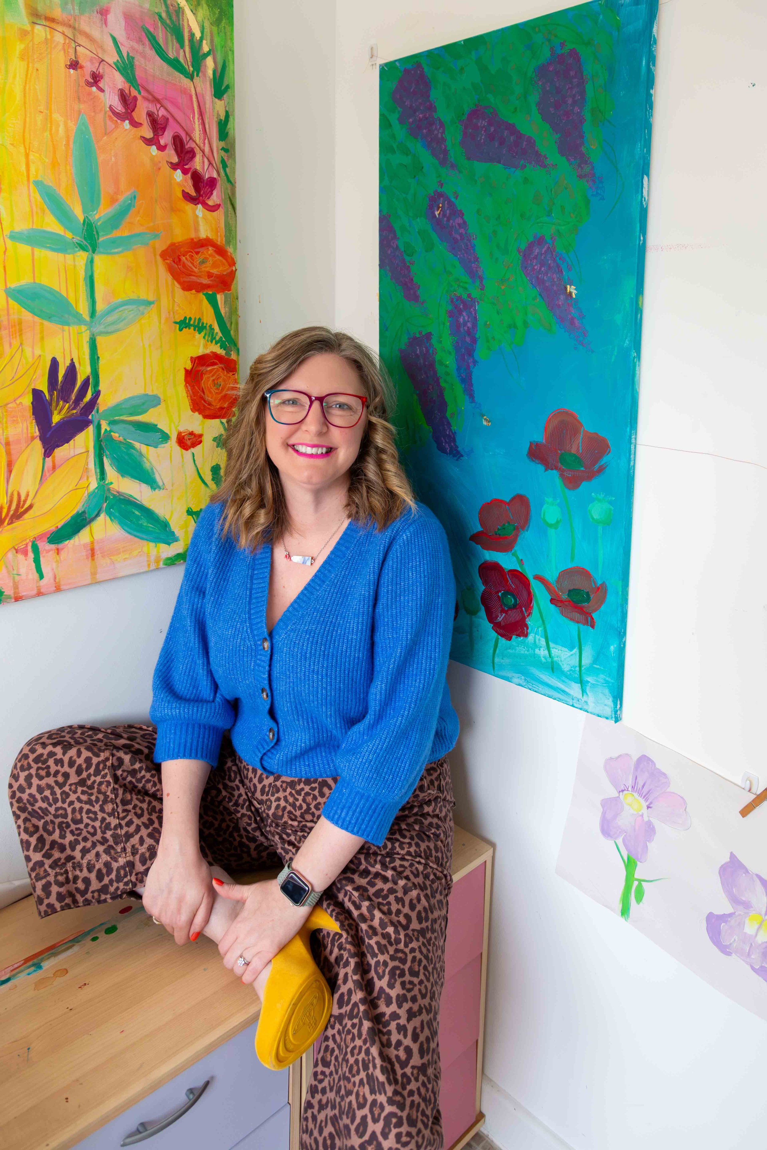 A woman with wavy light brown hair, glasses, and a big smile, sitting with legs crossed in a colorful art studio. She wears a blue cardigan, leopard print pants, and a smartwatch. Behind her are vibrant paintings of flowers and leaves on the walls.