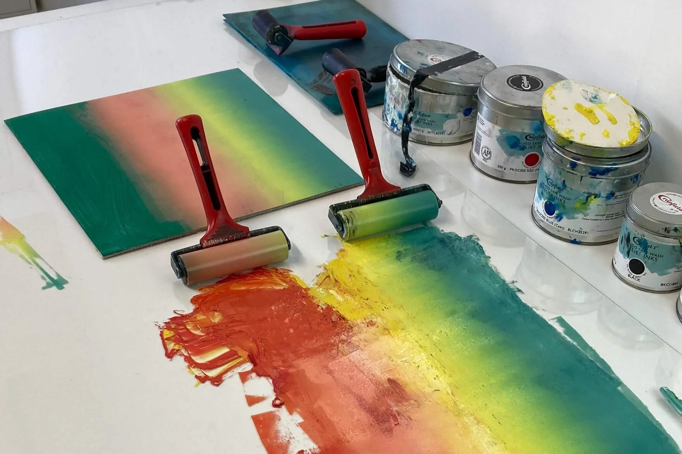 Artistic process with paint rollers covering paper with rainbow colors, surrounded by jars of paint and a palette knife.
