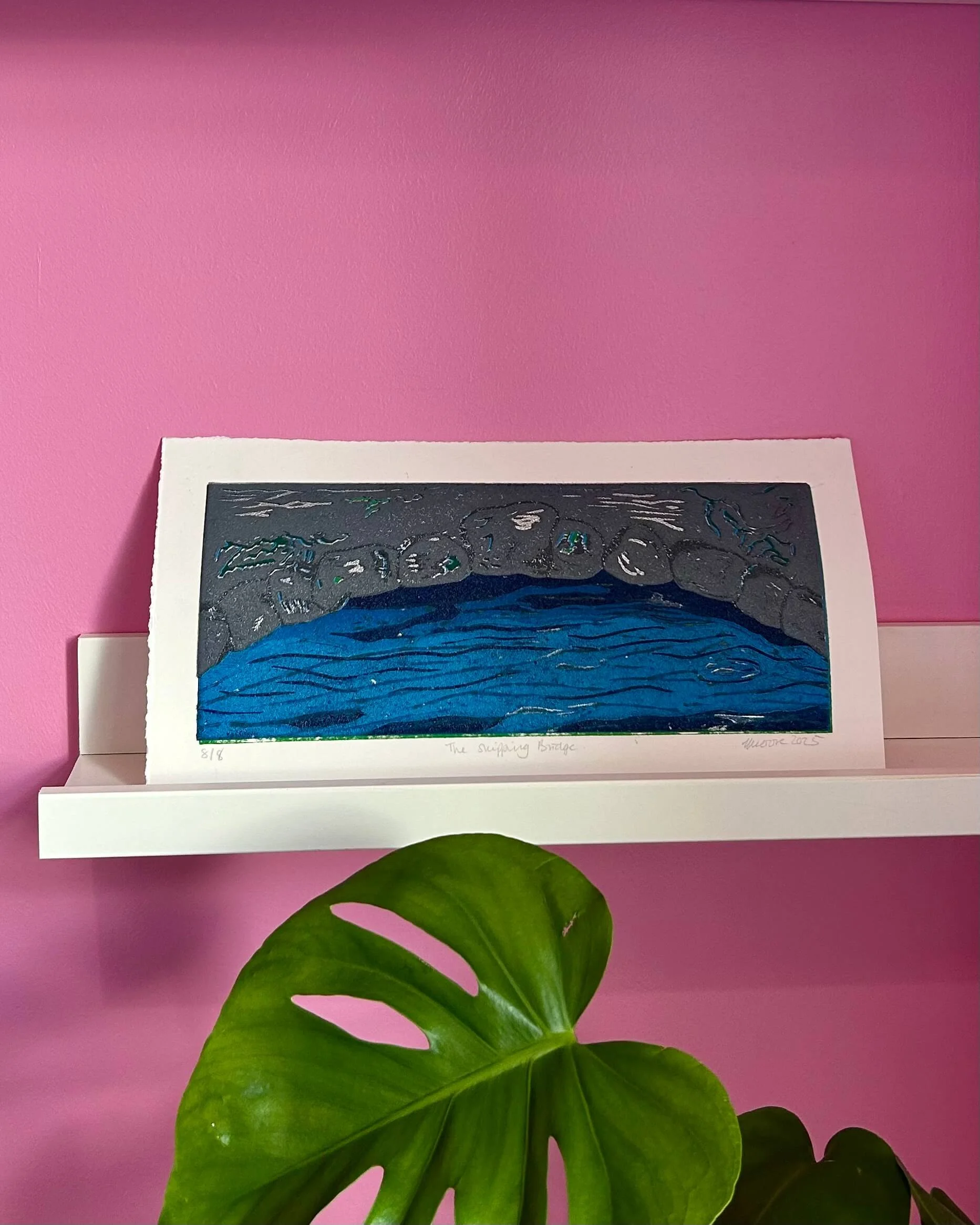 The Skipping Bridge original print on a white shelf in front of a pink wall - a lino reduction print of a silver grey stone bridge over a rippling blue river