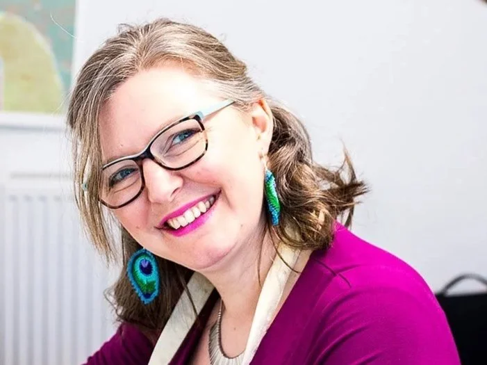 Artist Heather Moore sat in her studio wearing a purple dress and crocheted peacock feather earrings with statement pink lipstick - photo by Lauren McWIlliams