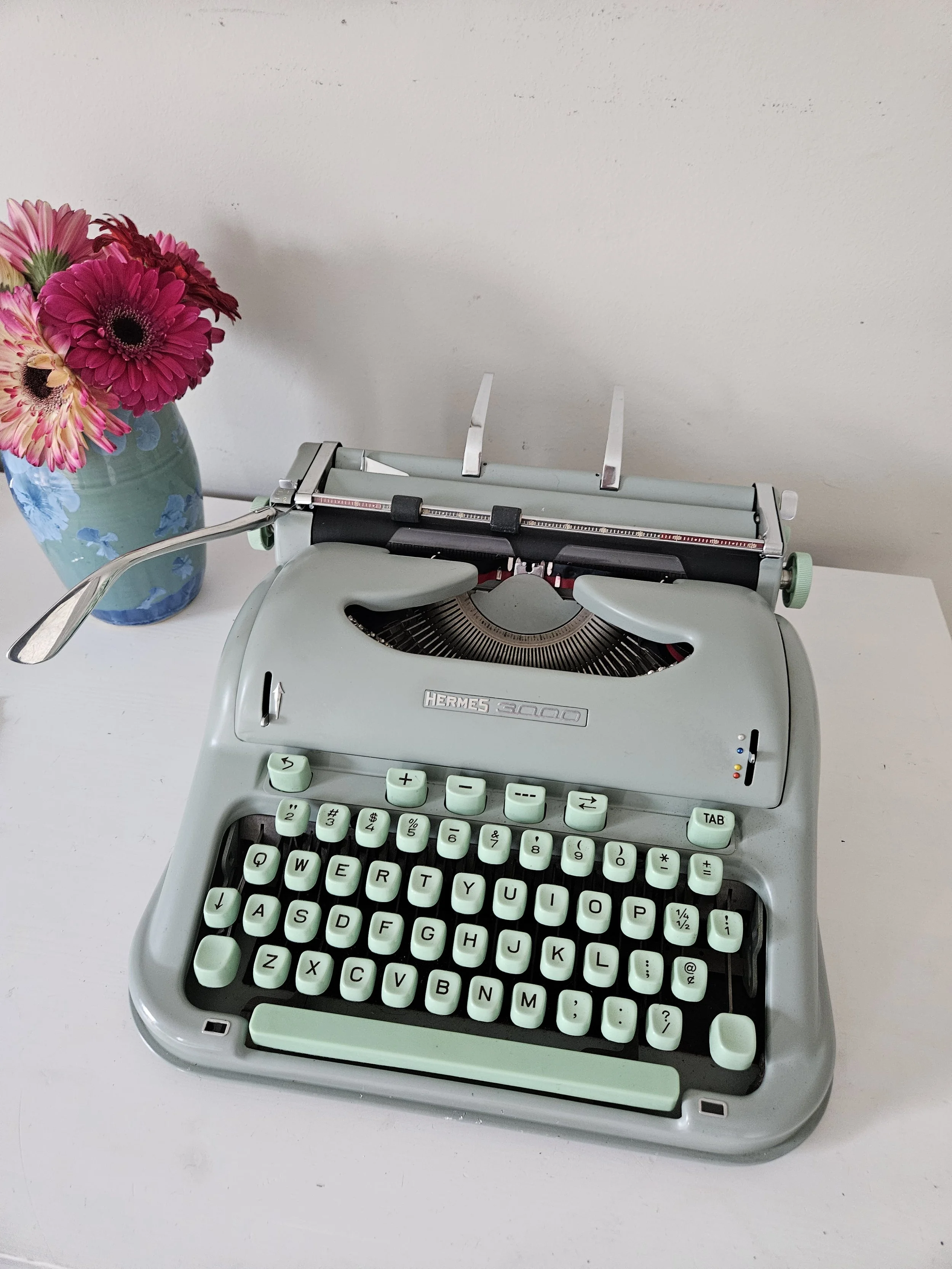 Restored Hermes 3000 typewriter in seafoam green with mint keys, beside a vase of pink gerbera daisies.