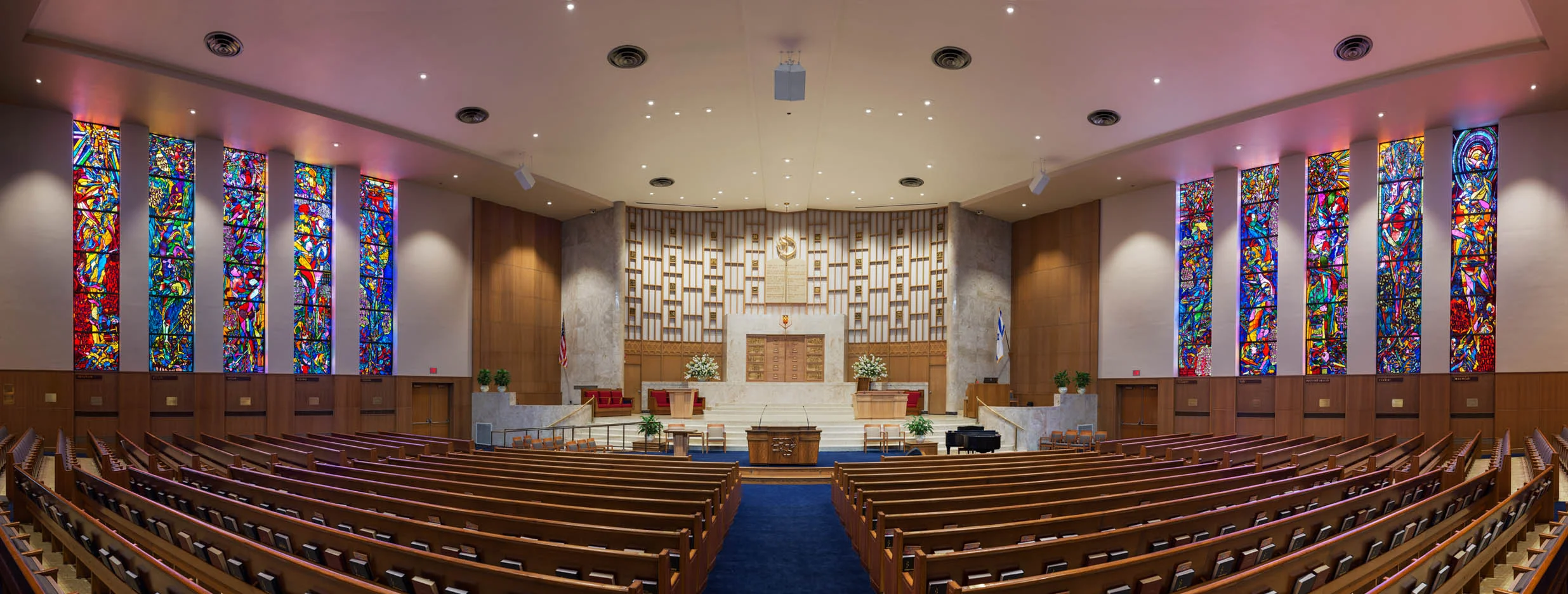 Reform Congregation Keneseth Israel — Sanctuary Renovation , Elkins Park, PA To meet the needs of an evolving congregation, Keneseth Israel turned to us to refresh the synagogue’s 1950s-era sanctuary in a manner respectful of its Modernist aesthetic. The main focus was the construction of a “low bimah” to provide a setting for smaller events in the large main sanctuary. By removing the first few pews, thoughtfully configuring a new low platform to include an ADA ramp, and creatively using energy-efficient lighting and digital controls, we worked to achieve a more intimate feeling for smaller groups of worshipers while retaining all of the beauty, drama and cherished traditions associated with the ark and broad upper bimah. Our team also assisted the synagogue’s design committee in selection of a new color scheme for carpet and fabrics to reupholster the existing auditorium seating and organ screen. New accent and overall sanctuary lighting levels were integrated with dimming controls to accommodate the addition of media projection screens. In addition to refurbishing the woodwork and finishes throughout the sanctuary, the team also restored the old upper bimah ceremonial furniture and designed new pieces for the low bimah.