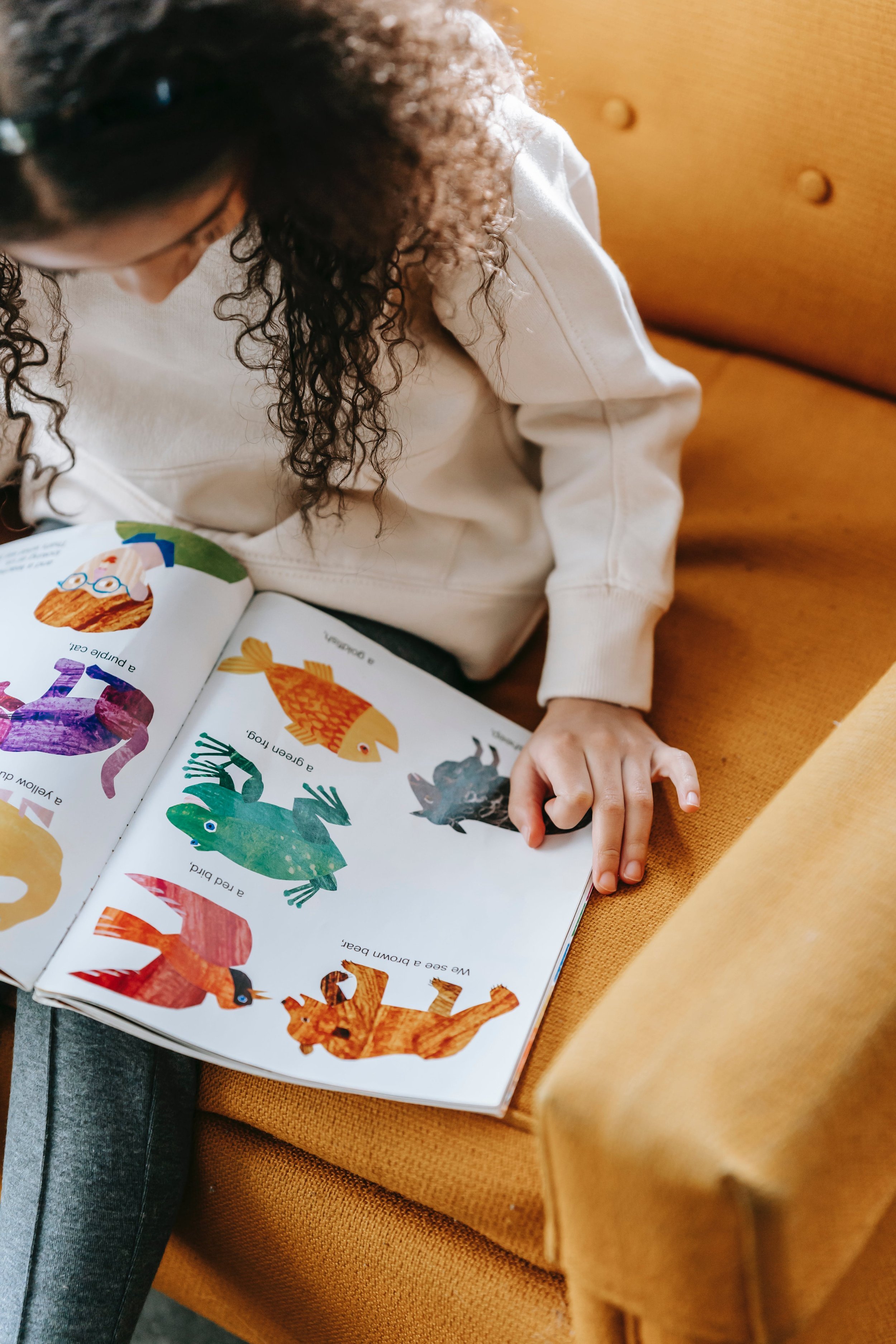 A young girl with curly brown hair, wearing a light beige sweatshirt, is sitting on an orange couch and reading a colorful picture book about animals.